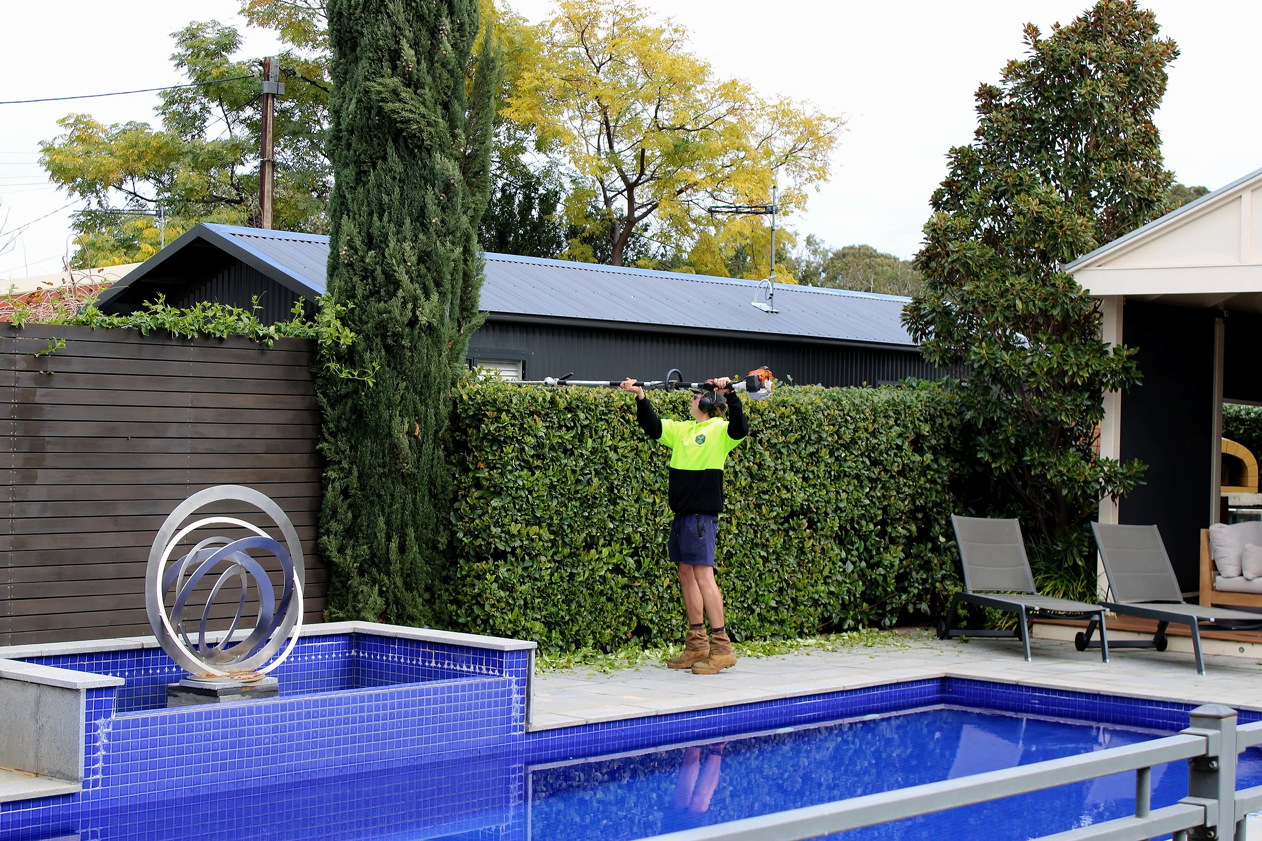 Person using a leaf blower to clear leaves from a backyard hedge near a swimming pool with blue tiles. Backyard features outdoor furniture, a wooden privacy fence, tall trees, and a modern house roof.