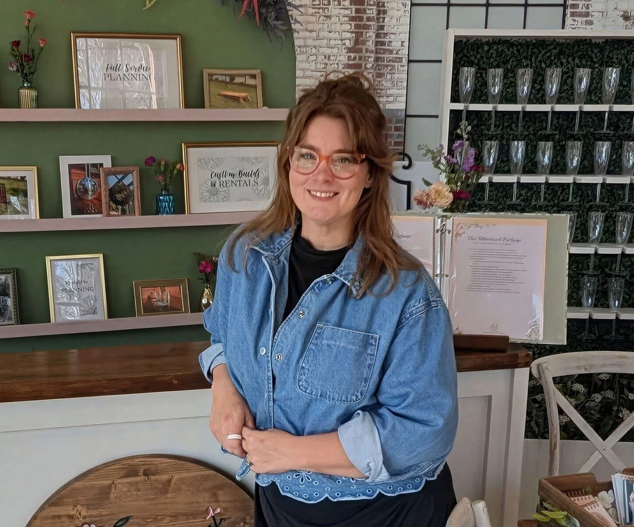 A woman with glasses, wearing a denim jacket over a black shirt, smiling in a cozy, decorated interior with shelves of picture frames, flowers, and glassware.