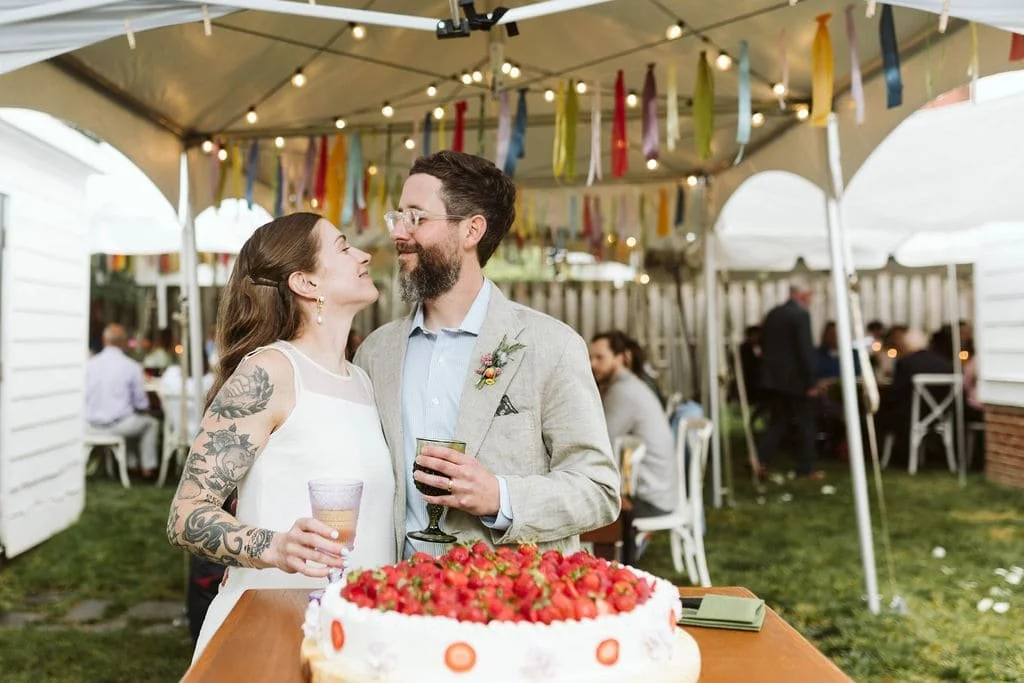 A couple at a wedding reception, standing under a tent with colorful ribbons and string lights, with a cake topped with strawberries on a table in front of them. The woman has tattoos on her arm, and the man is holding a drink while looking at her, both smiling.