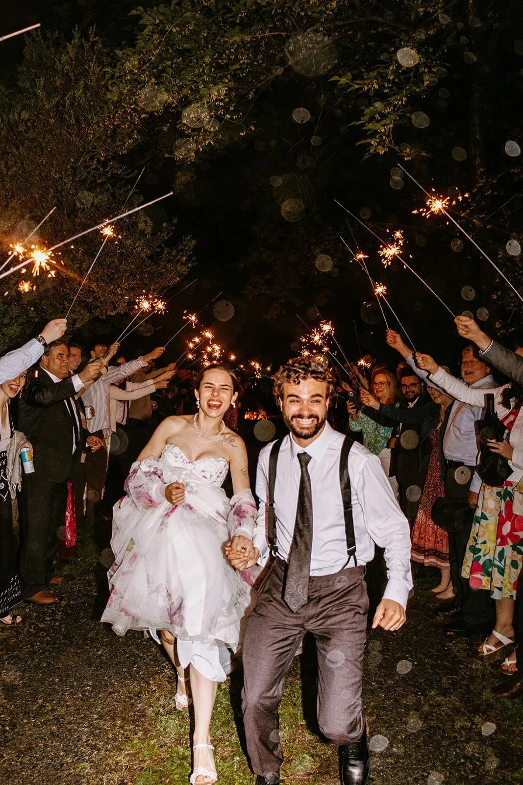 A newly married couple smiling and holding hands, walking through a crowd at night, with people holding sparklers and celebrating.
