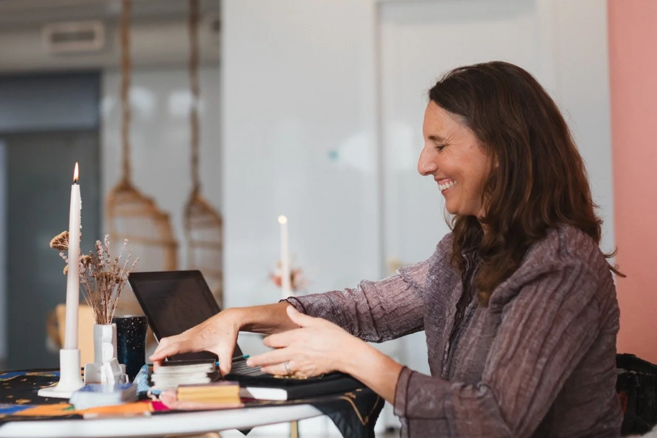 A woman with brown hair is smiling and reaching into a card deck on a table decorated with candles, flowers, and books.