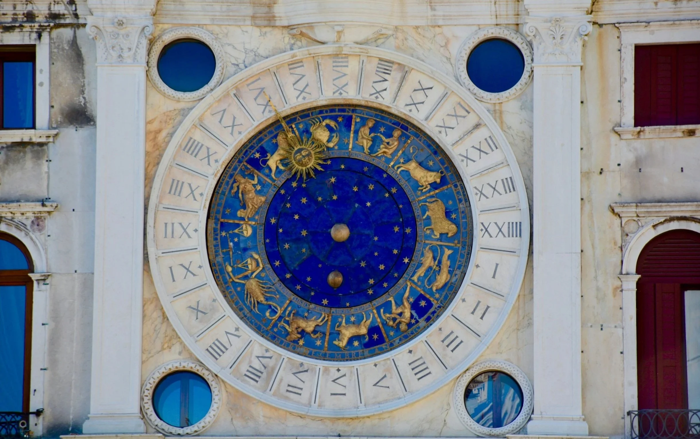 Close-up of a historic clock tower with zodiac signs and Roman numerals, mounted on an aged stone building with windows.