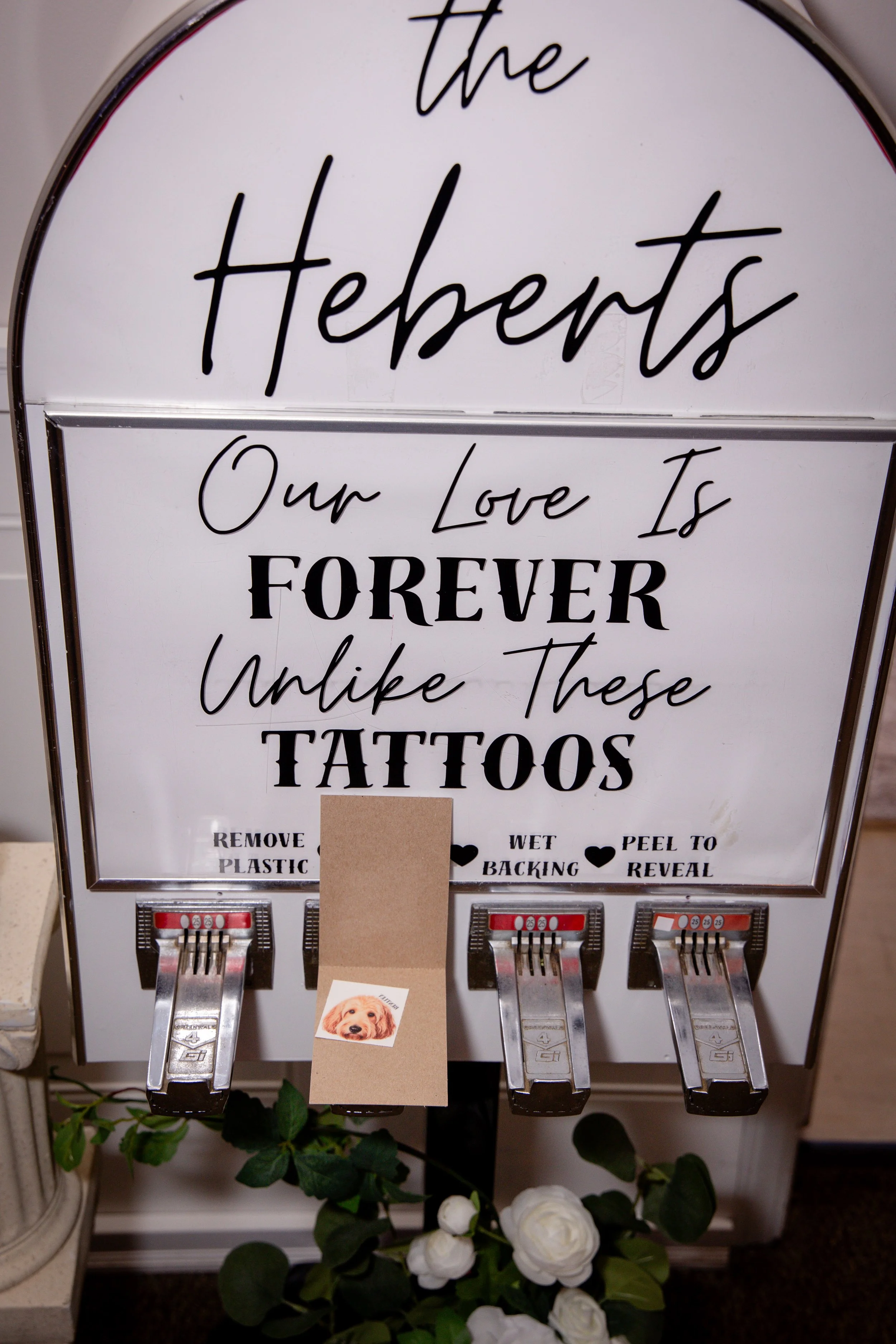 A driftwood-style move sign with black cursive and block lettering that reads, "the Heberts Our Love Is Forever" and instructions for tattoo removal. Below the sign are three photo booth style detectors.