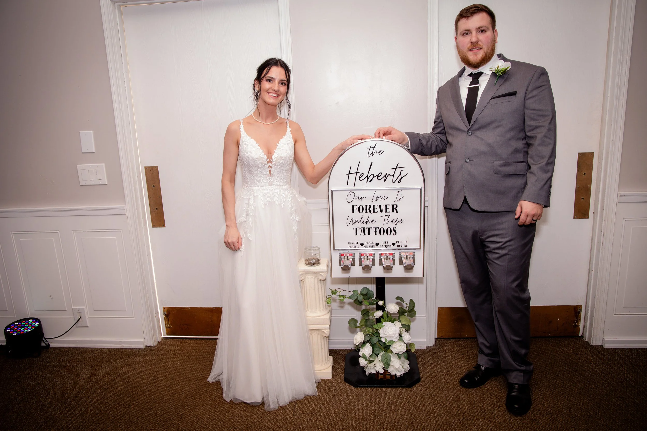 A bride and groom standing next to a sign at their wedding reception. The bride is wearing a white bridal gown and pearl necklace, and the groom is in a gray suit with a black tie and boutonniere. The sign reads 'the Heberts, Our Love Is Forever, Unless These Tattoos,' and lists instructions for removing or covering tattoos.