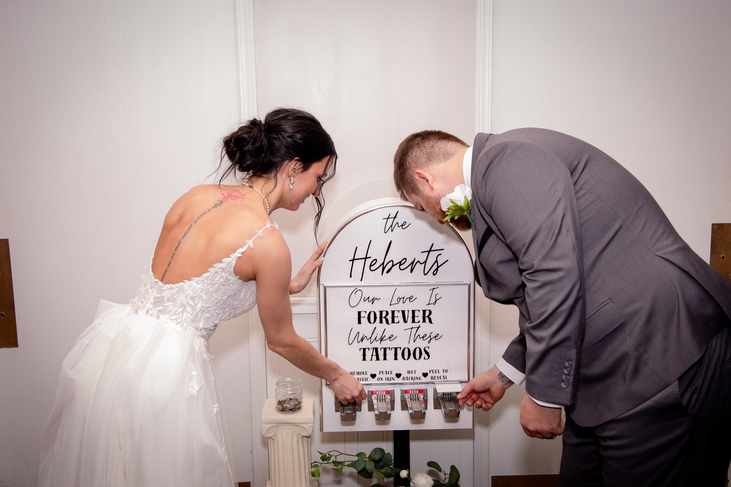 A bride and groom are bowing their heads towards a sign that reads, 'the Heberts, Our Love Is Forever, Unlike These Tattoos,' at a wedding celebration. The bride is wearing a white wedding dress and the groom is in a gray suit.