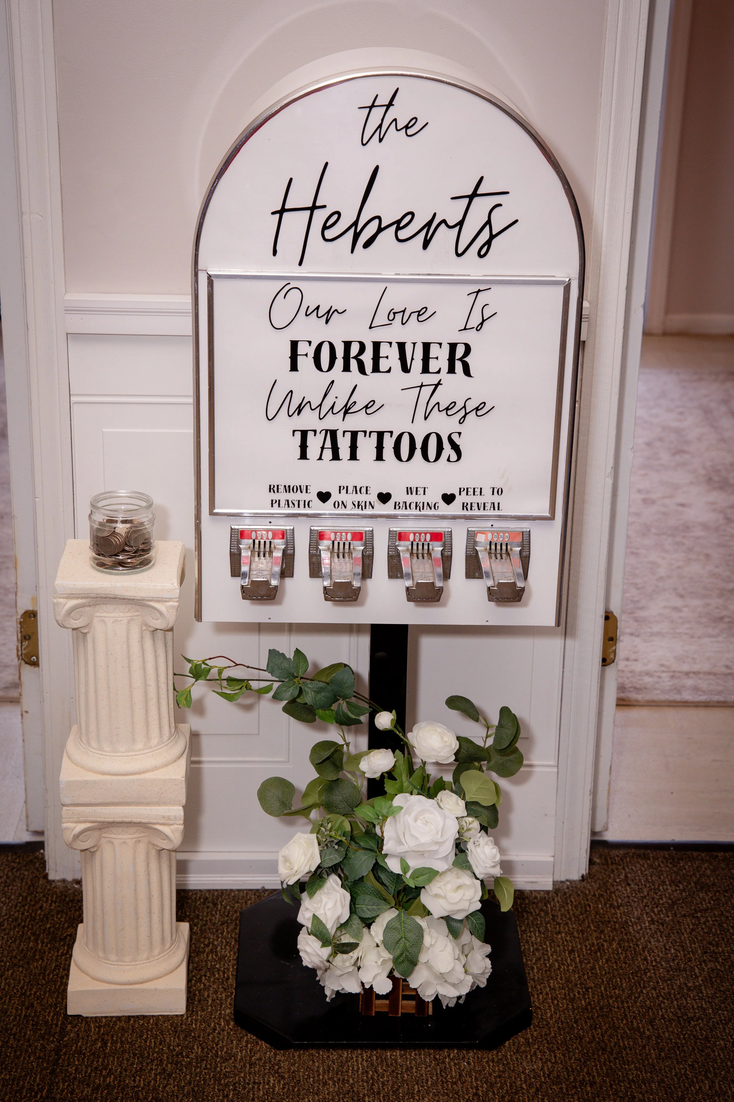 A decorative sign that reads 'The Hebarts' with the message 'Our Love Is Forever Unlike These Tattoos'. The sign is white with black text and is displayed on a stand, with a small jar of coins on a pedestal, next to artificial white roses and greenery on a black base. The background includes a white wall and beige carpet.