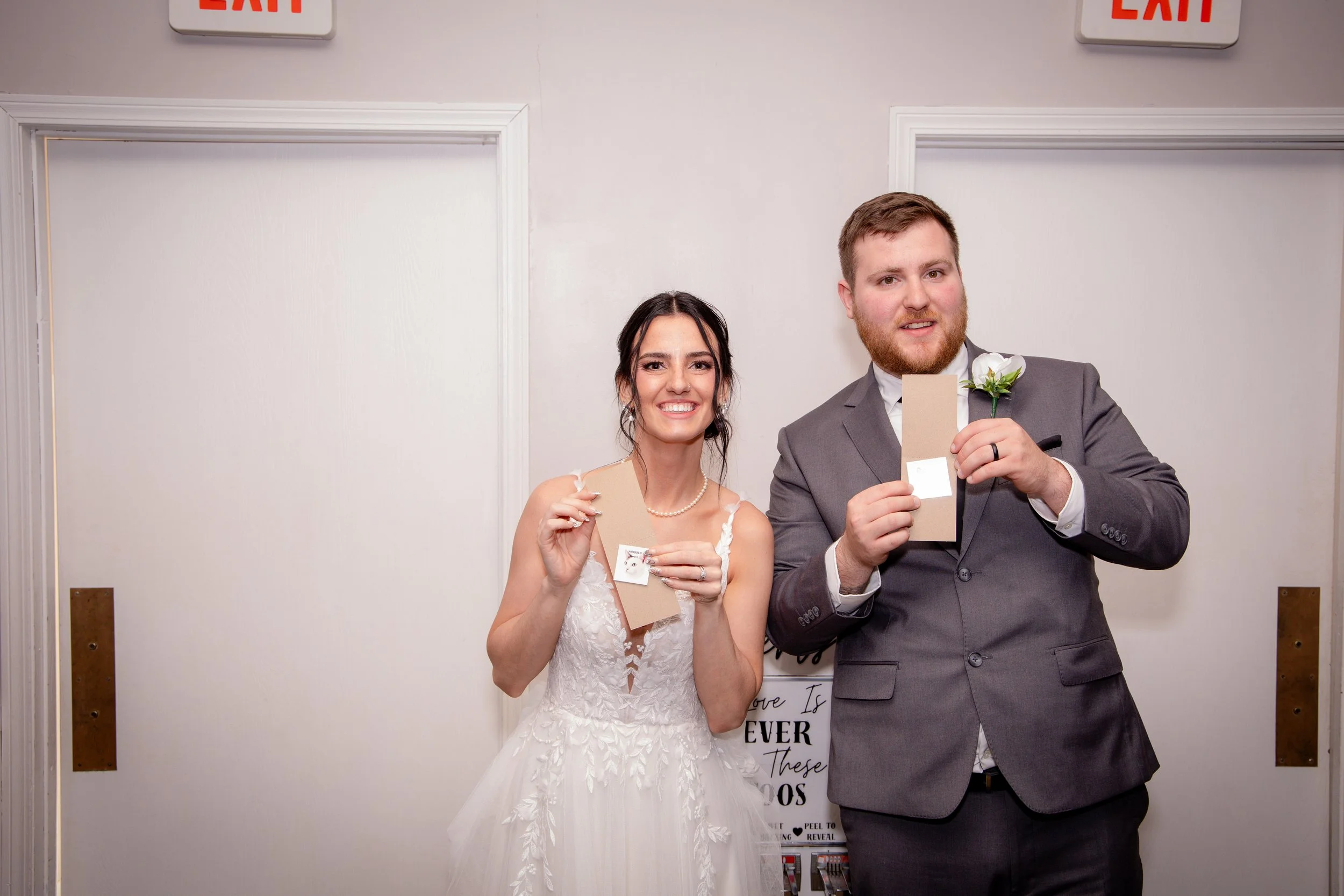 A bride and groom holding beige cards and smiling at the camera in front of a plain wall with exit signs. The bride is wearing a white wedding dress with lace details and a pearl necklace. The groom is dressed in a grey suit with a white shirt, black tie, and a white flower boutonniere.