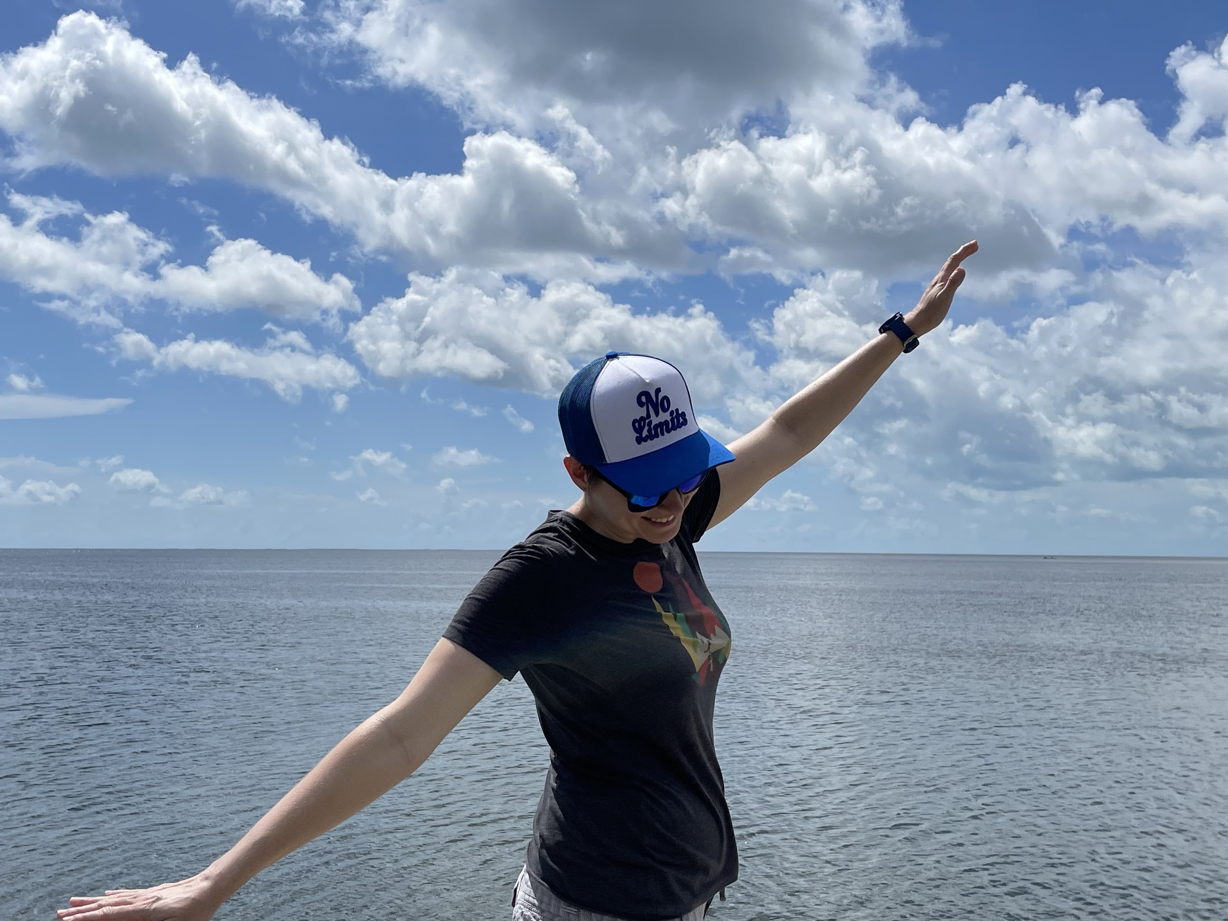 Coach Elena standing in front of the ocean on a cloudy day with her arms outstretched as if flying, wearing a hat that reads "No Limits"