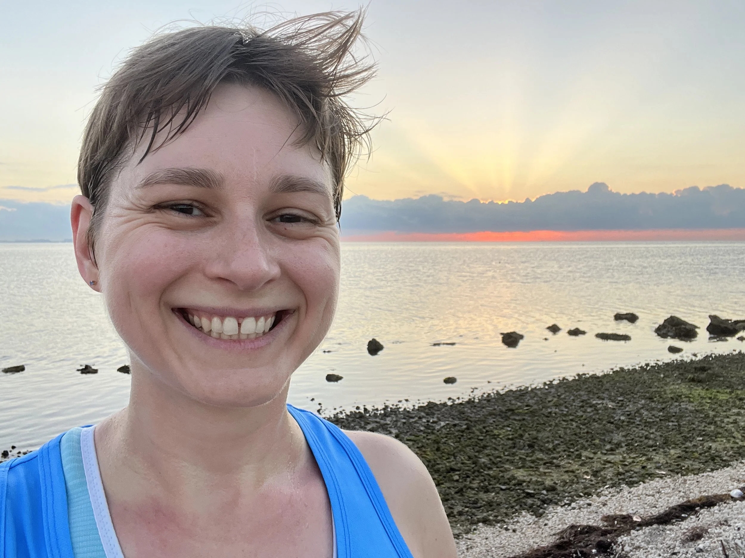 Coach Elena smiling at the camera in front of an ocean sunrise during a run.