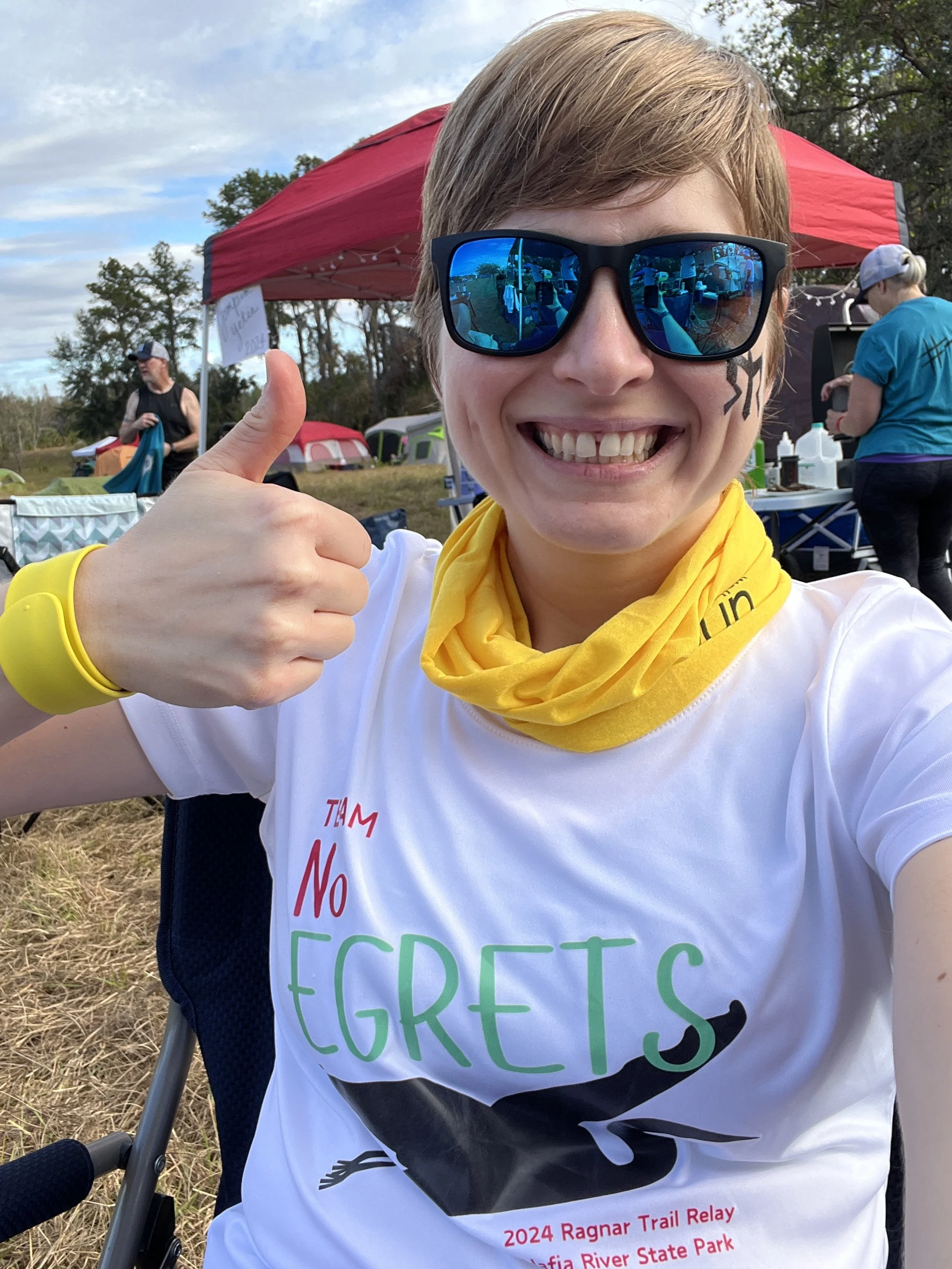 Coach Elena at a Ragnar Trail campground with a big smile and thumbs up wearing a "Team No Egrets" t-shirt
