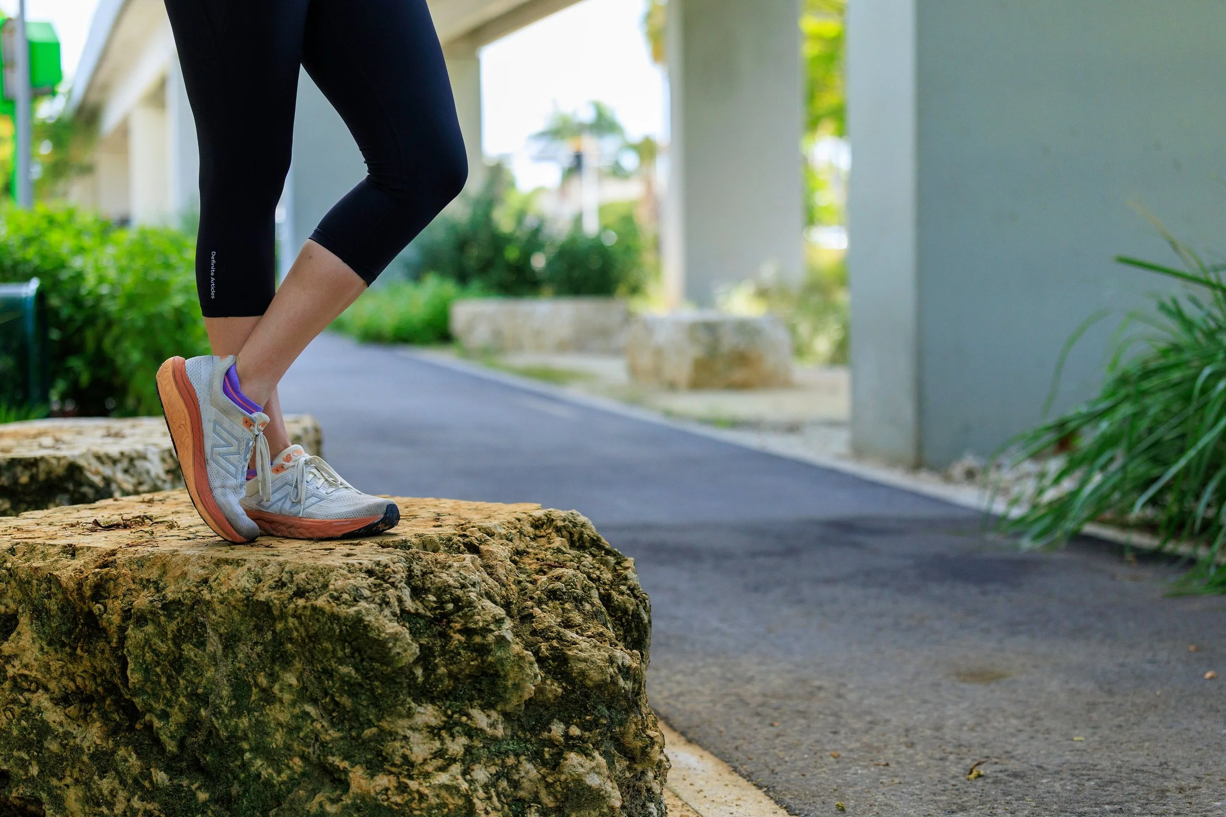 Close-up of a person wearing sneakers, standing on a rock in an outdoor setting with greenery and a pathway in the background.