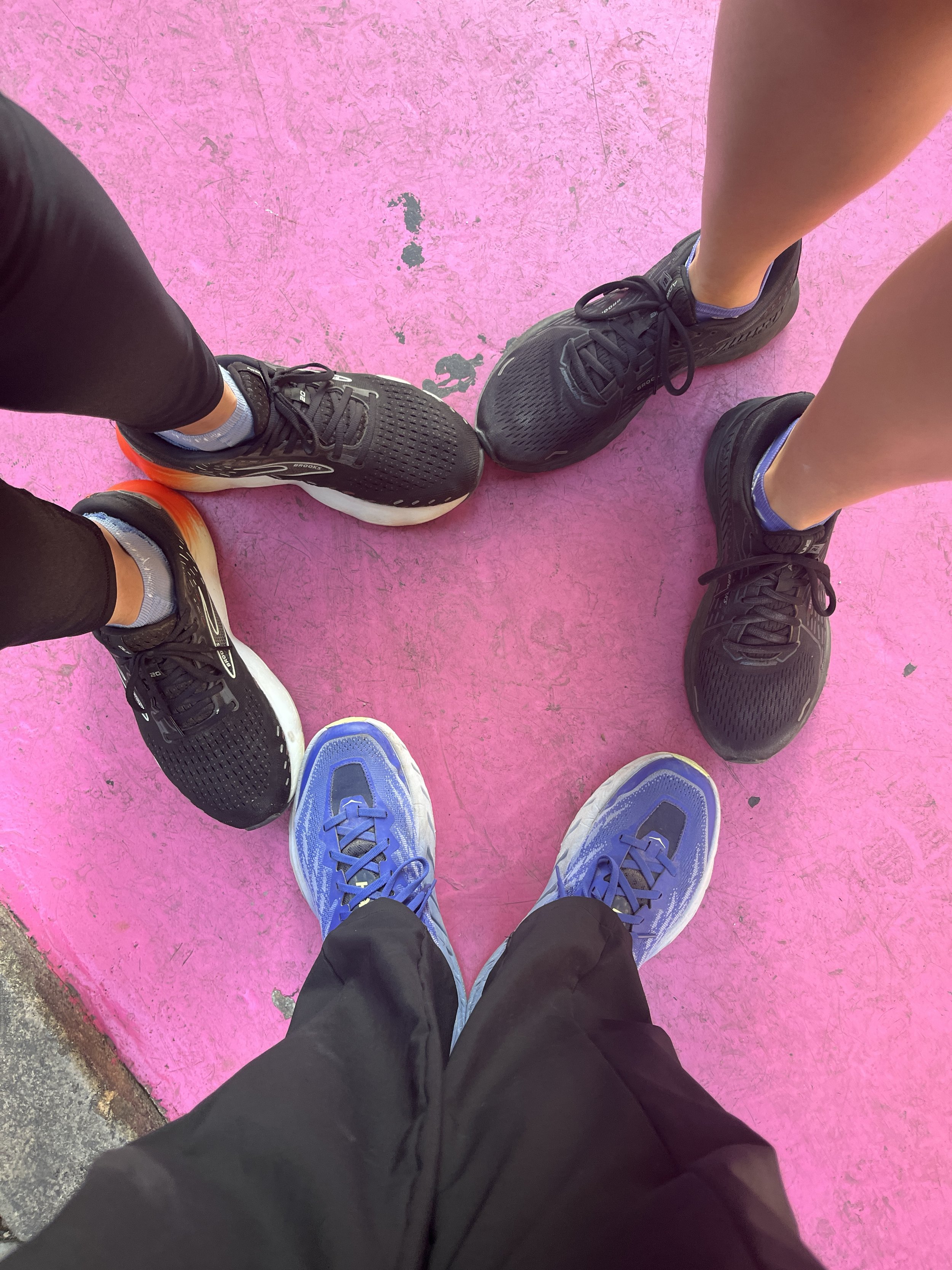 View of three people's running shoes and ankles as they stand together to create a heart with their shoes over a pink sidewalk