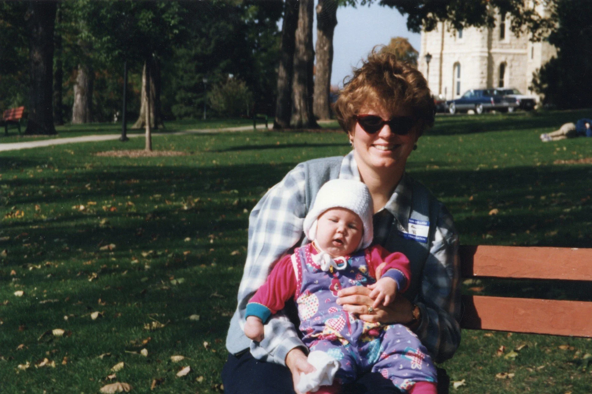 A woman with short curly hair, wearing sunglasses and a plaid shirt, sitting on a park bench holding a baby girl dressed in colorful clothing and a white knit hat. There are trees, grass, and a historic building in the background, and a person lying on the grass.
