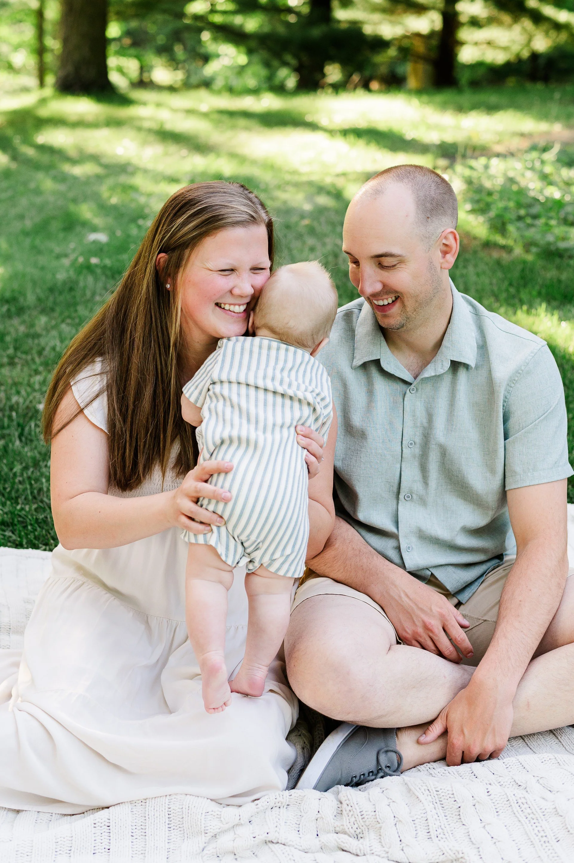 A family of three, a woman, a man, and a baby, enjoying time together outdoors on a grassy area under trees. The woman is holding the baby while smiling, the man is sitting beside them, also smiling. They are dressed casually and appear happy and engaged with each other.