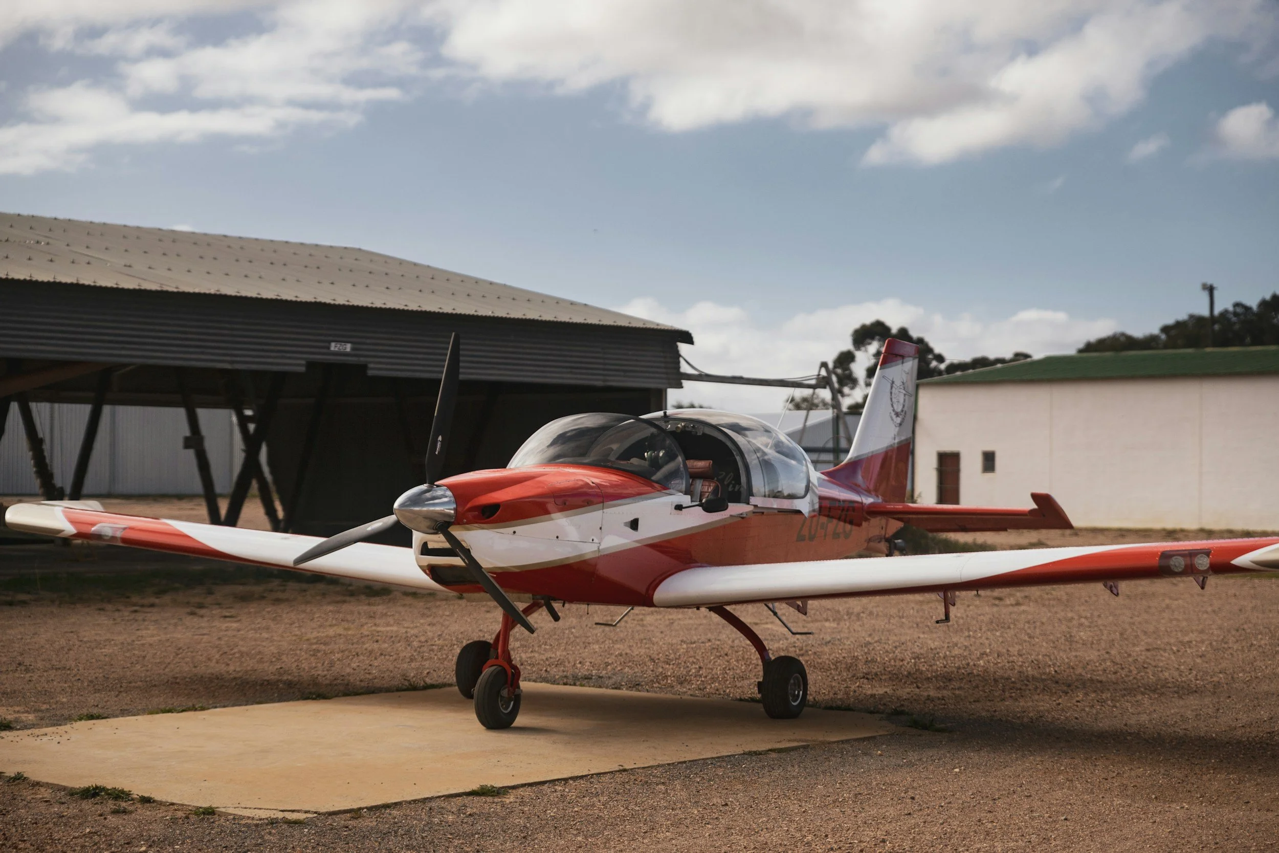 Red and white small aircraft on tarmac with buildings and trees in background under cloudy sky.