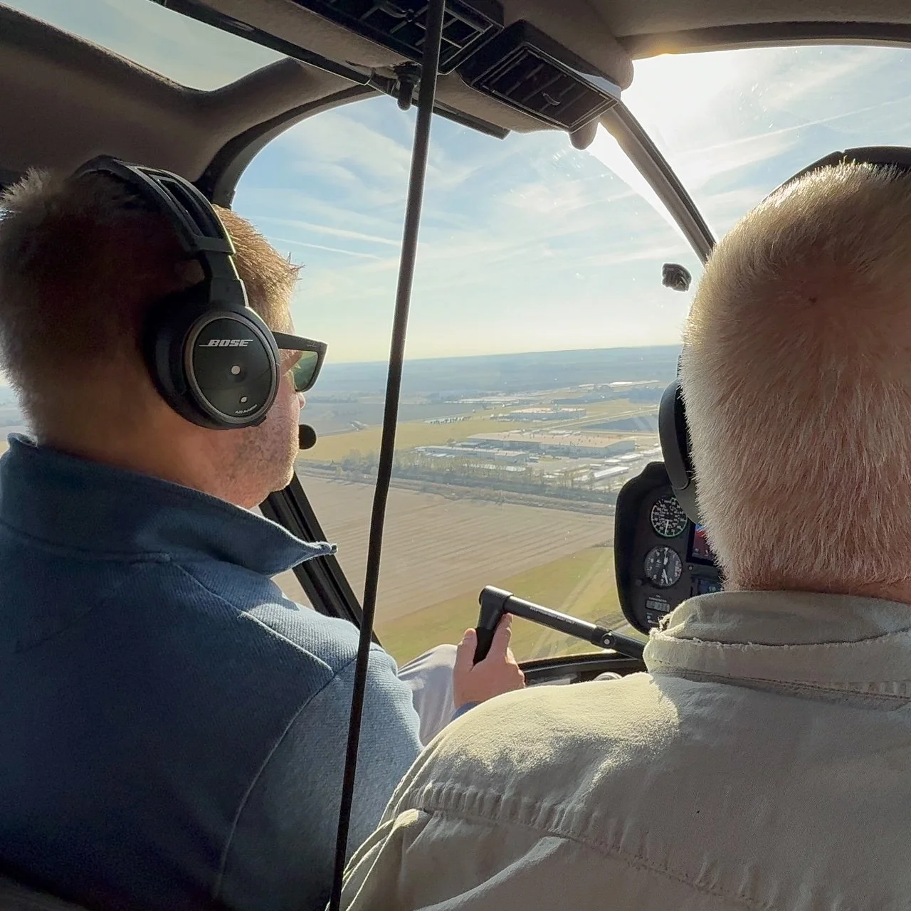 Two men flying in a helicopter over farmland, wearing headsets, with one man holding the controls and the landscape visible through the helicopter's window.