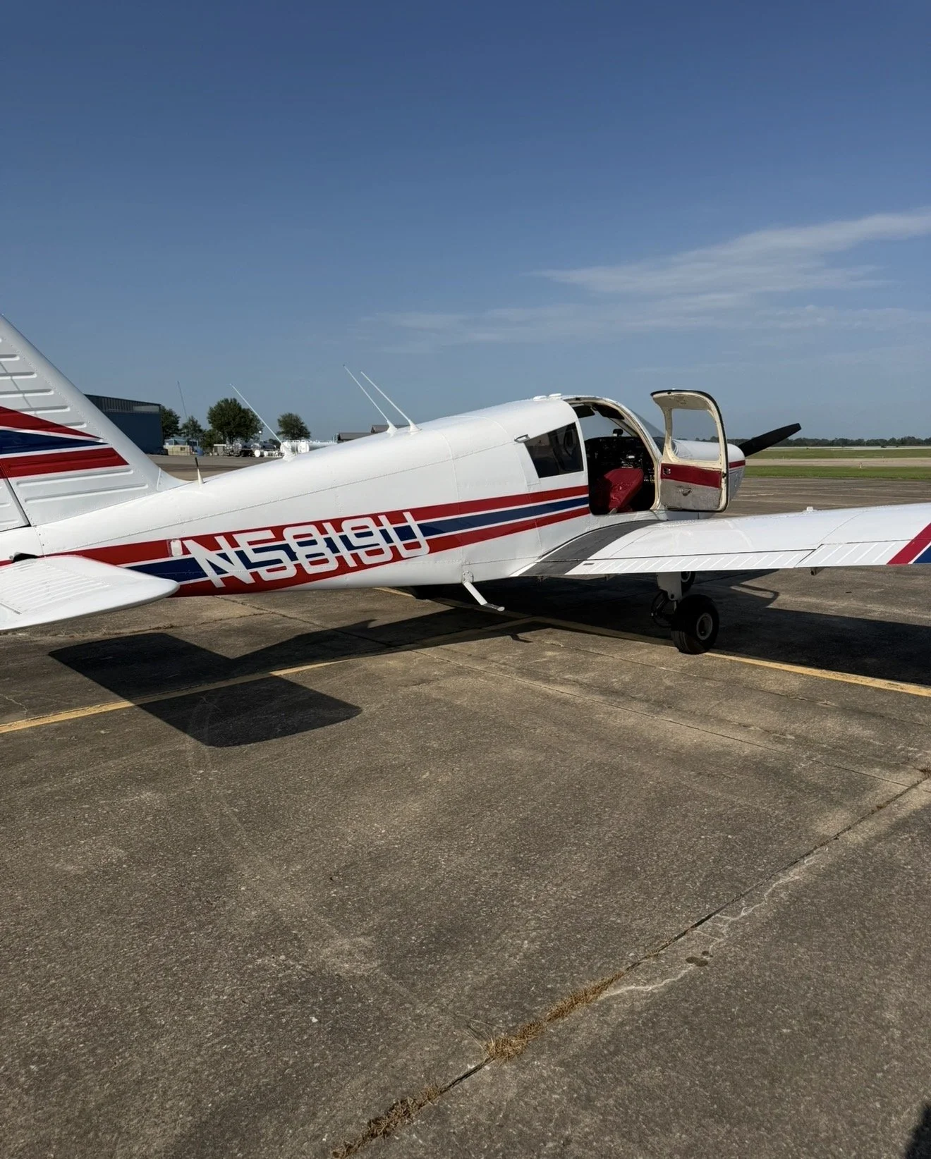 Small white airplane with red and blue stripes on the side, parked on the tarmac at an airfield under a clear blue sky.