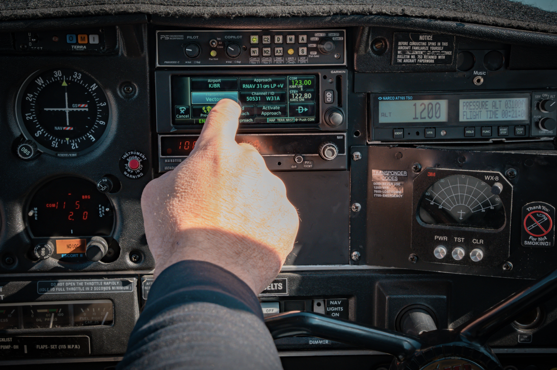 Interior view of an aircraft cockpit with a pilot's hand adjusting controls on the central panel, surrounded by various gauges, screens, and switches.
