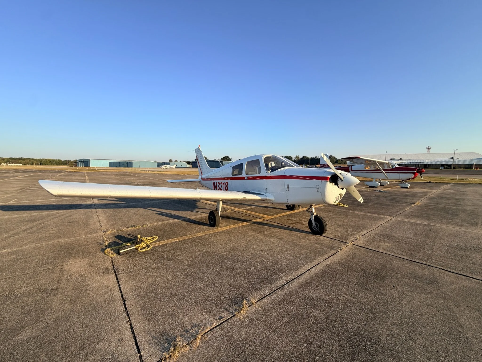 Small white and red private airplane parked on an airport tarmac with hangars and a clear blue sky in the background.