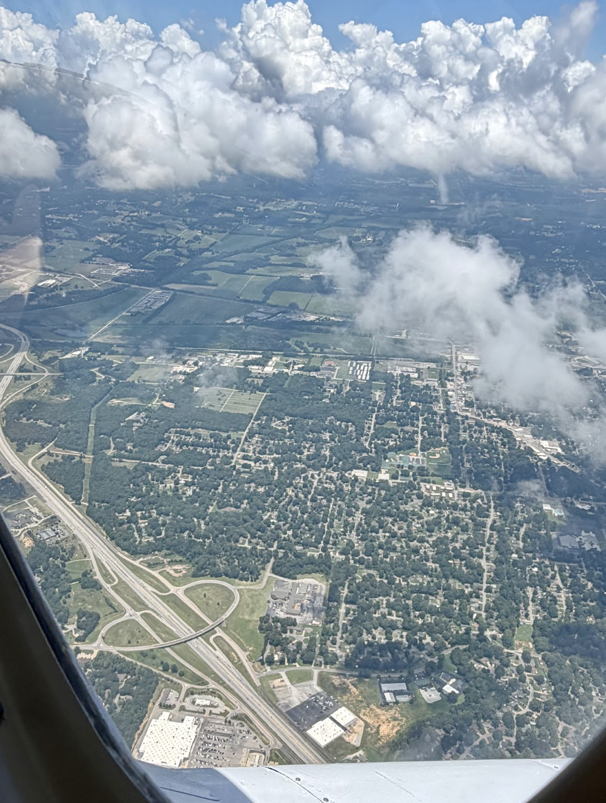 Aerial view of a landscape with green fields, roads, and scattered buildings seen through an airplane window with clouds overhead.