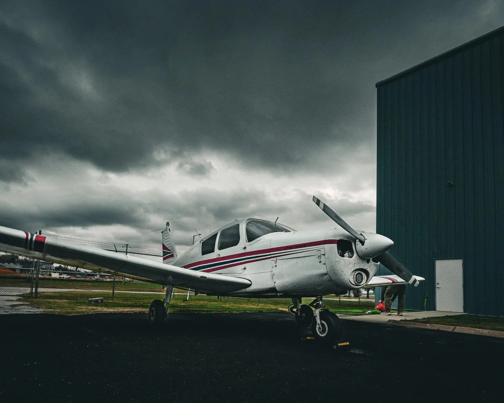 Small white airplane with red and black stripes parked outside a grey hangar under a dark, cloudy sky. A person is standing near the hangar.