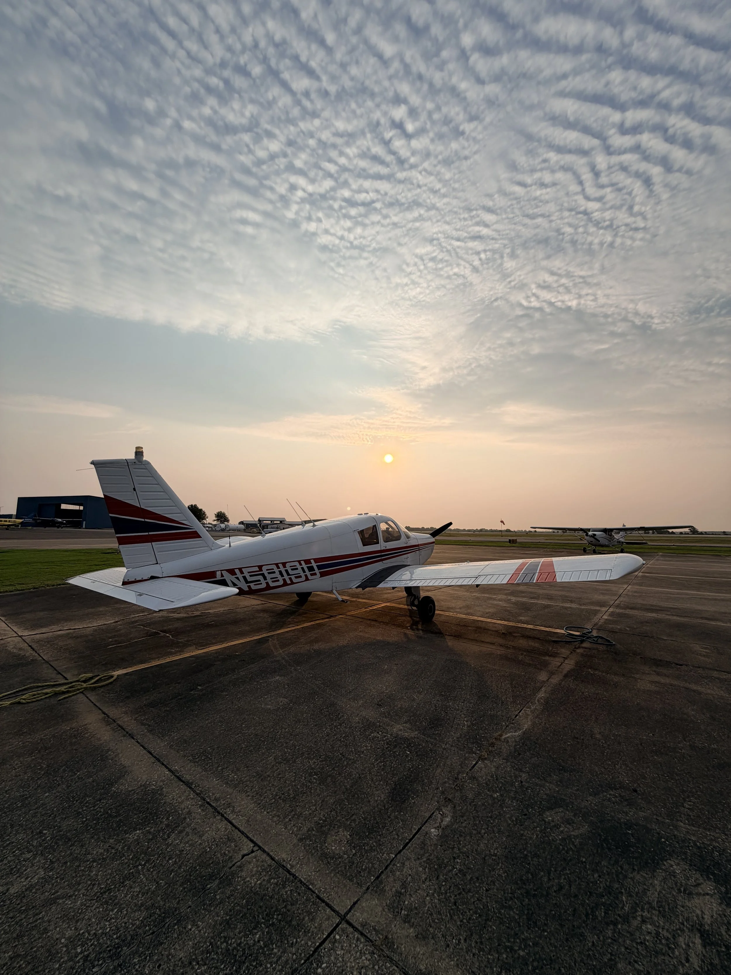 Small white airplane with red and black stripes on the runway at sunset, with other aircraft and hangars in the background under a partly cloudy sky.
