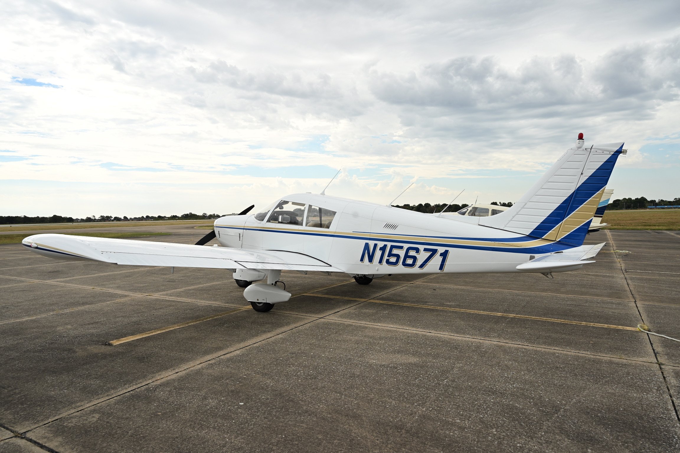 Small white airplane with blue and gold stripes parked on a tarmac, with a cloudy sky above and another airplane in the background.