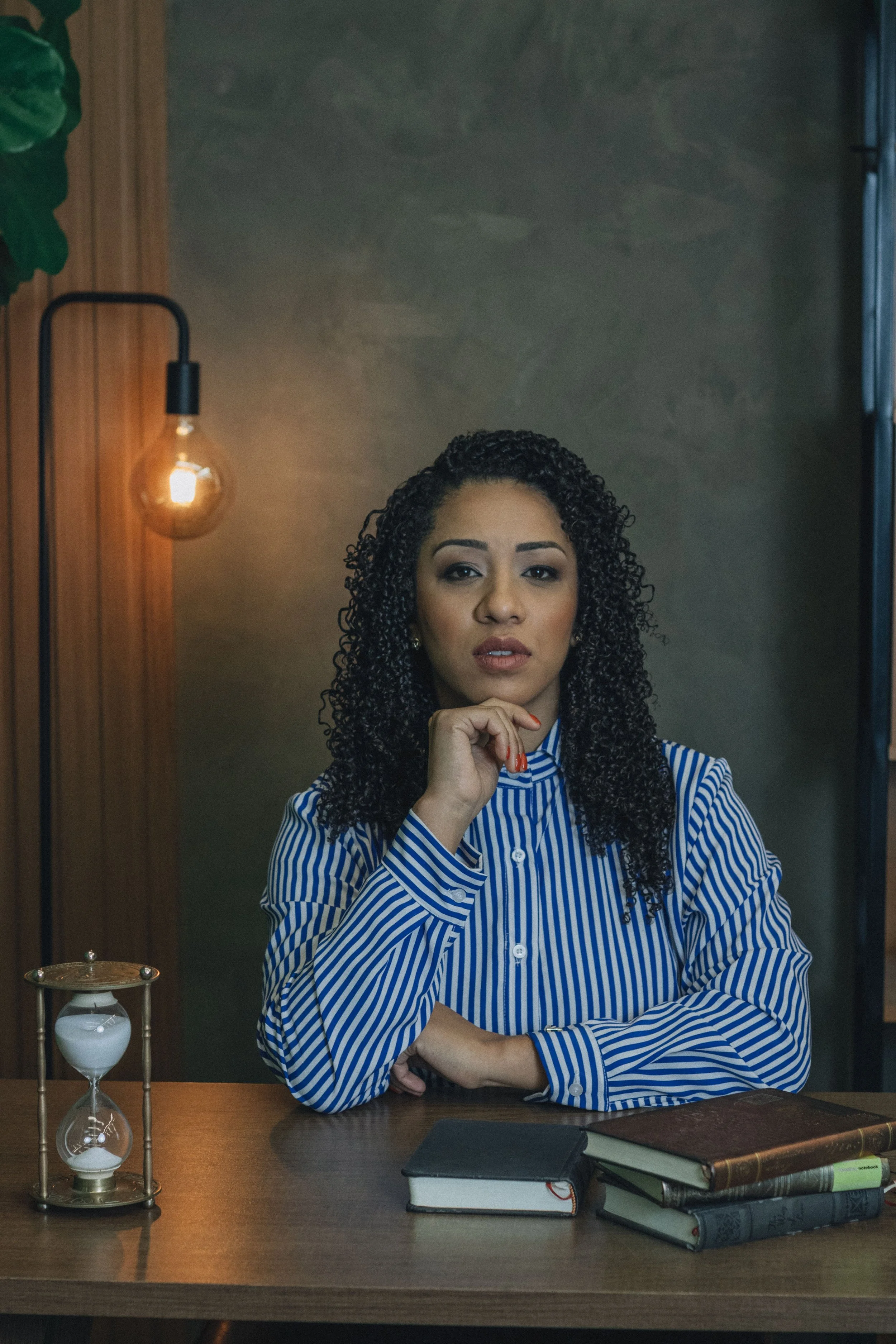 a woman seated at a desk with a dark background, an a lamp illuminating her face to the left. There is a small hour glass and books on her work desk. Her face is pensive. Her right hand is below her chin, posed as if she is thinking.
