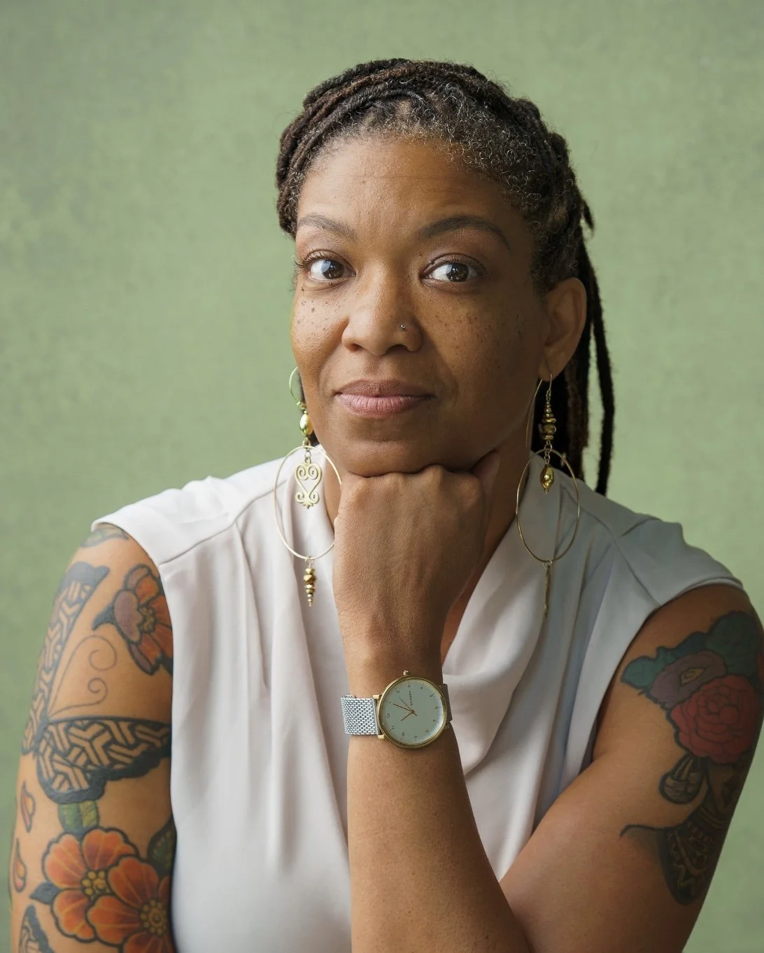 A woman with tattoos on her arms, wearing a white sleeveless top, earrings, a watch, and a thoughtful expression, resting her chin on her hand against a green background.