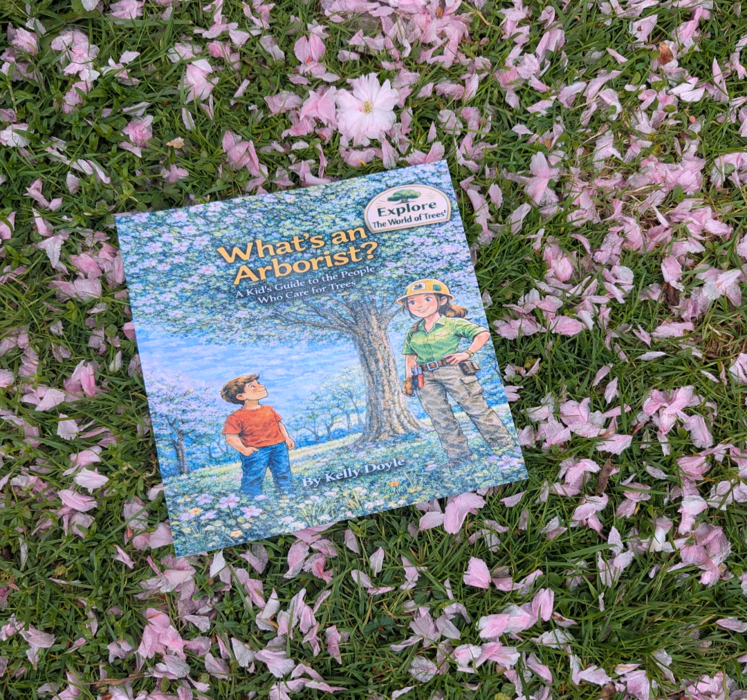 A children's book titled "What's an Arborist?" resting on pink flower petals and green grass.