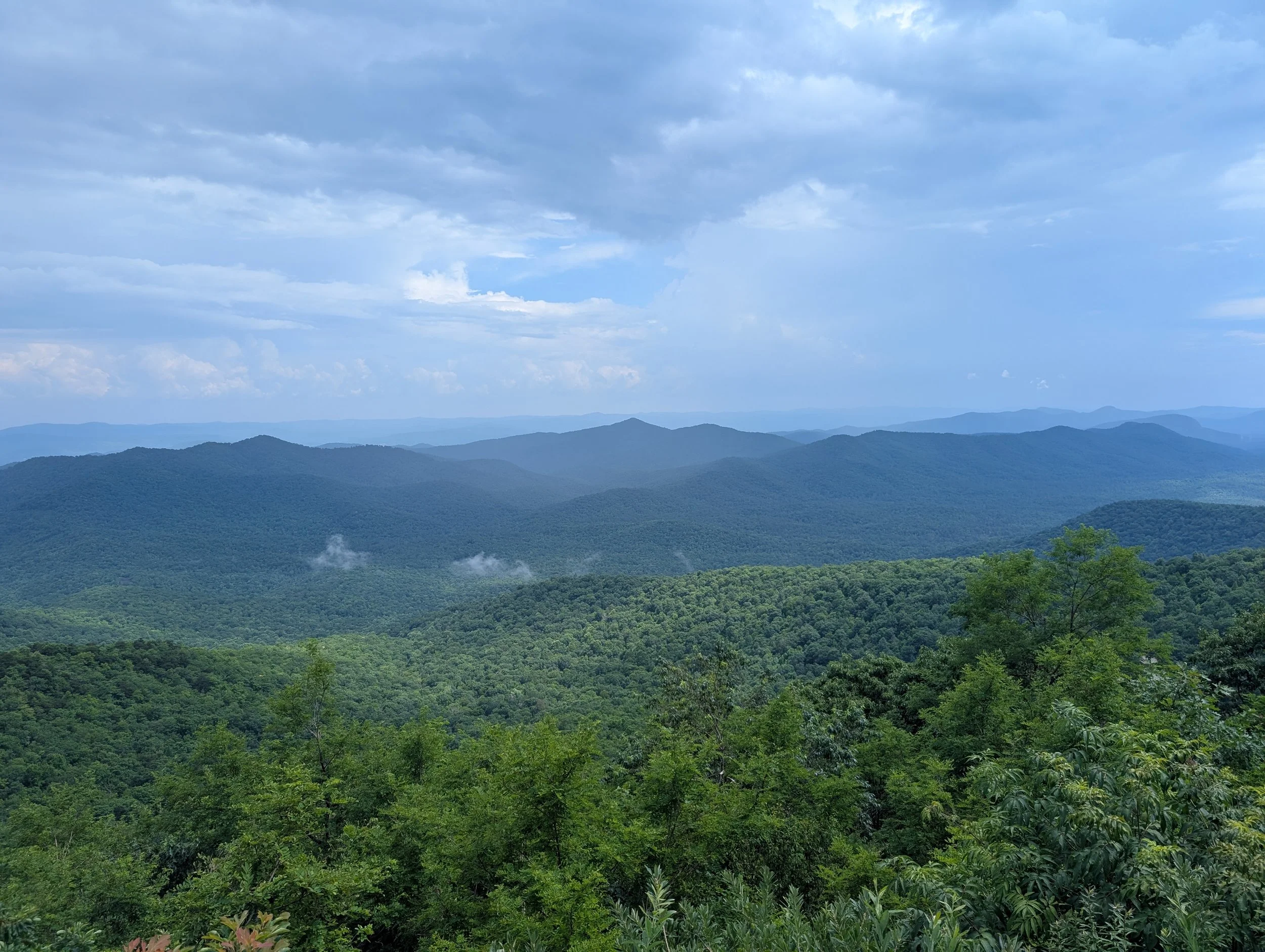 View of rolling green mountains with dense forest, under a cloudy sky.