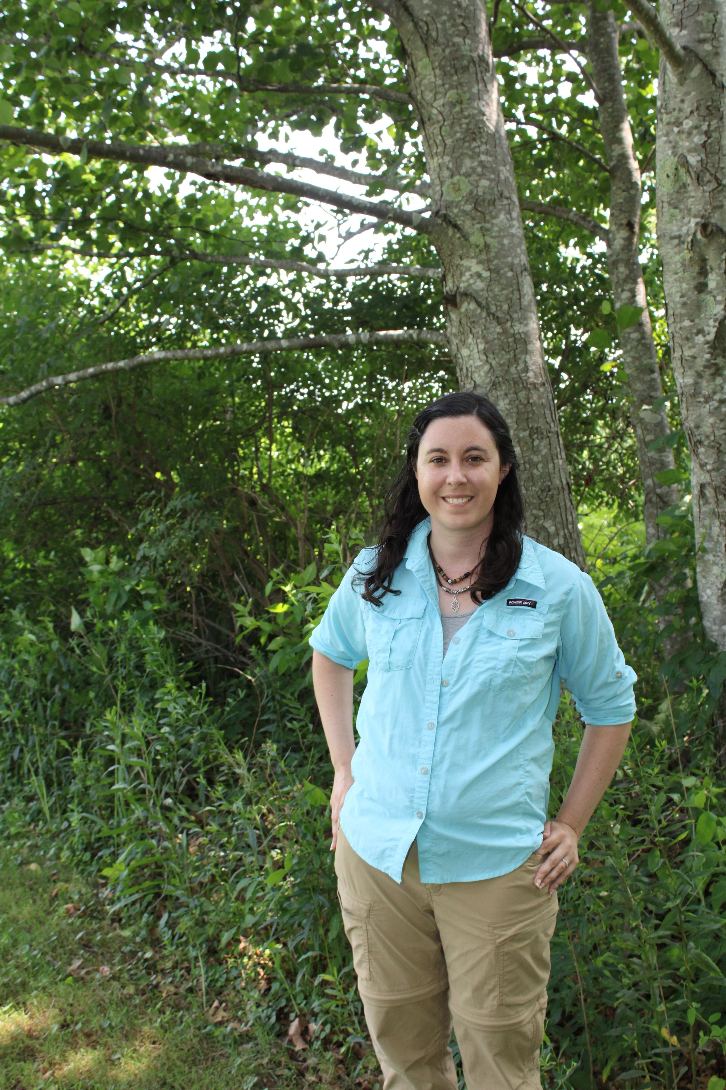 A woman with long dark hair smiling outdoors in front of trees and greenery. She is wearing a light blue button-up shirt and khaki pants.