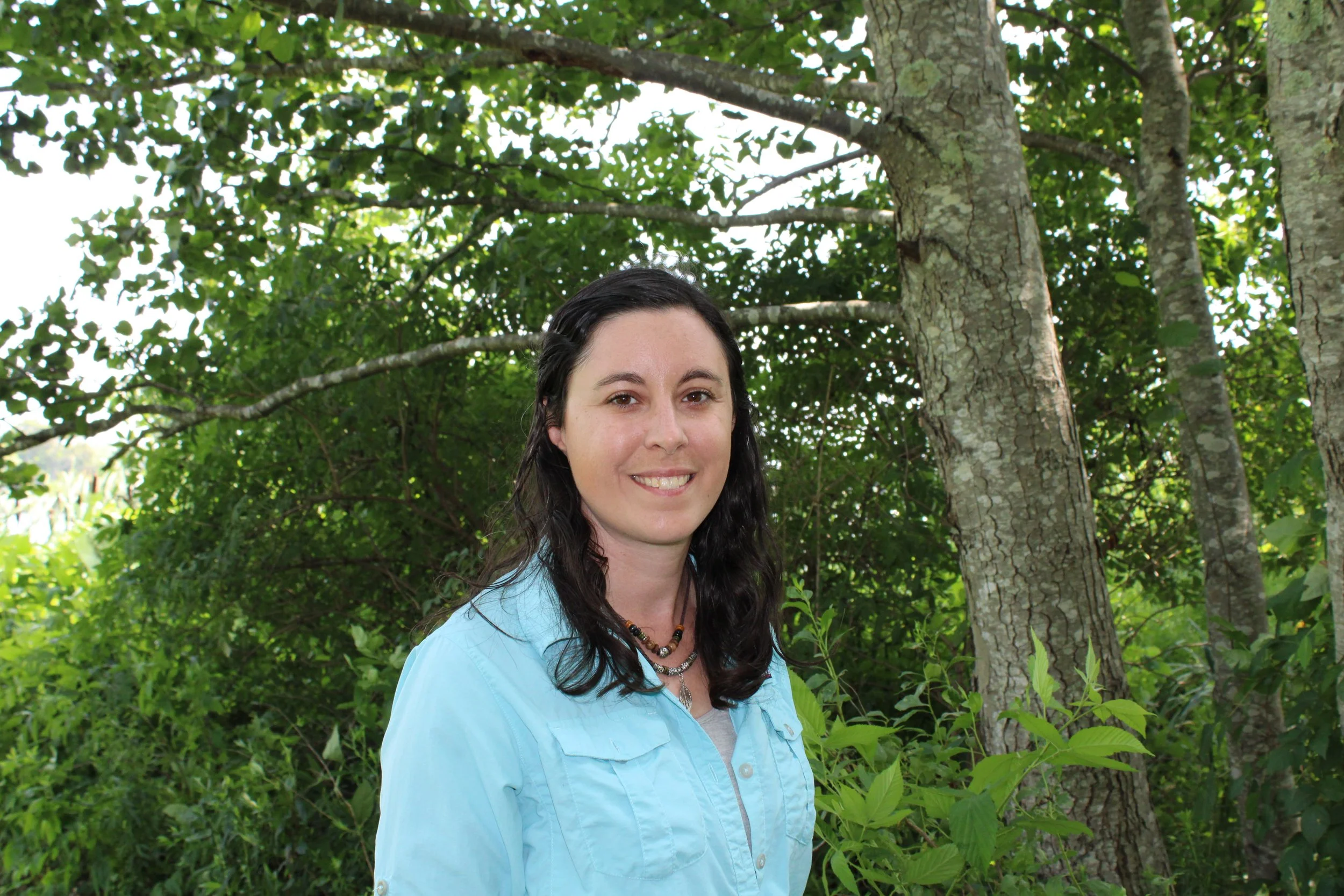 A woman with long dark hair and a light blue shirt standing in a lush green forest with trees and dense foliage.
