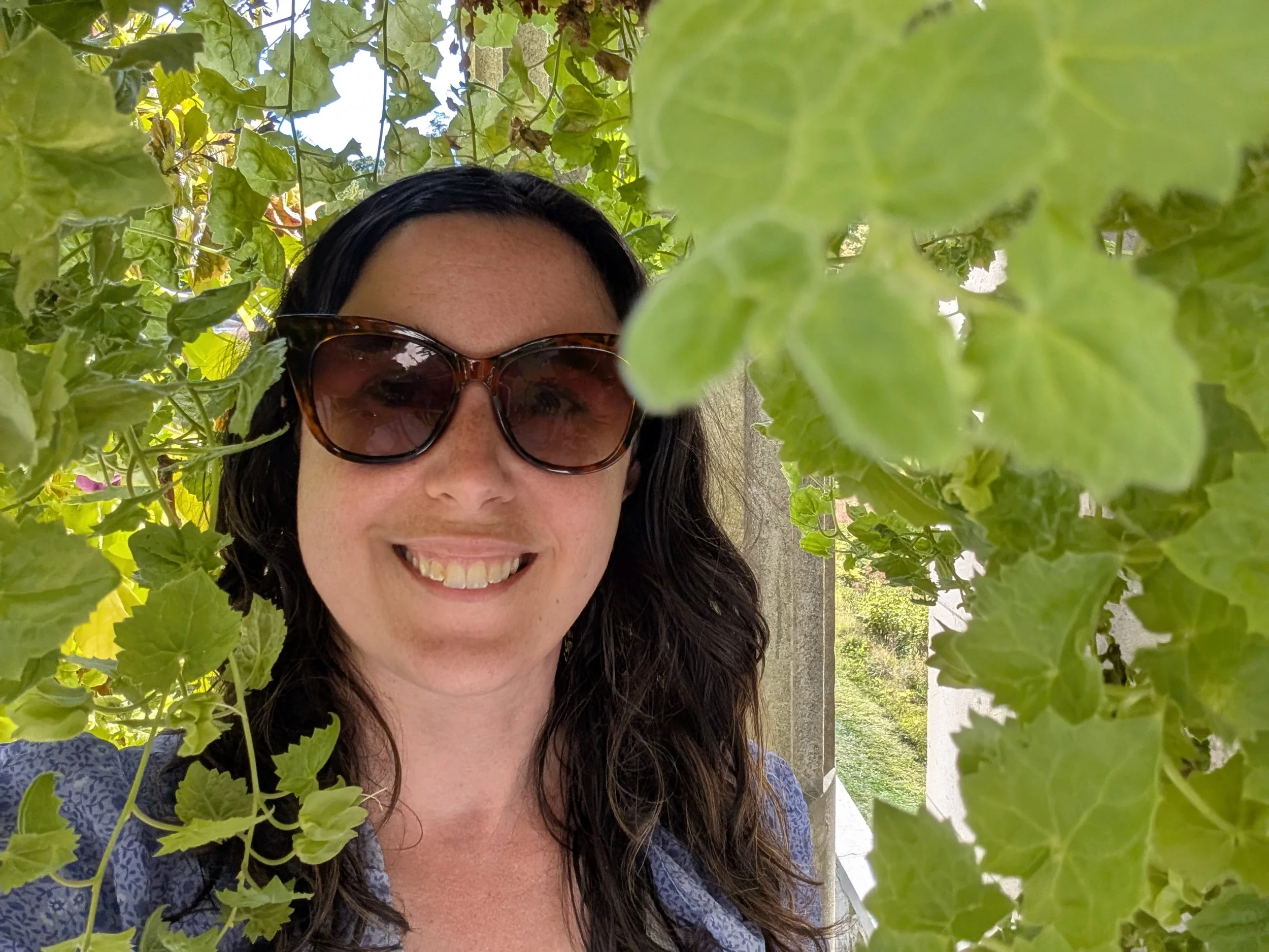 A woman with dark hair, wearing sunglasses, smiling as she stands among green leafy plants outdoors during daytime.