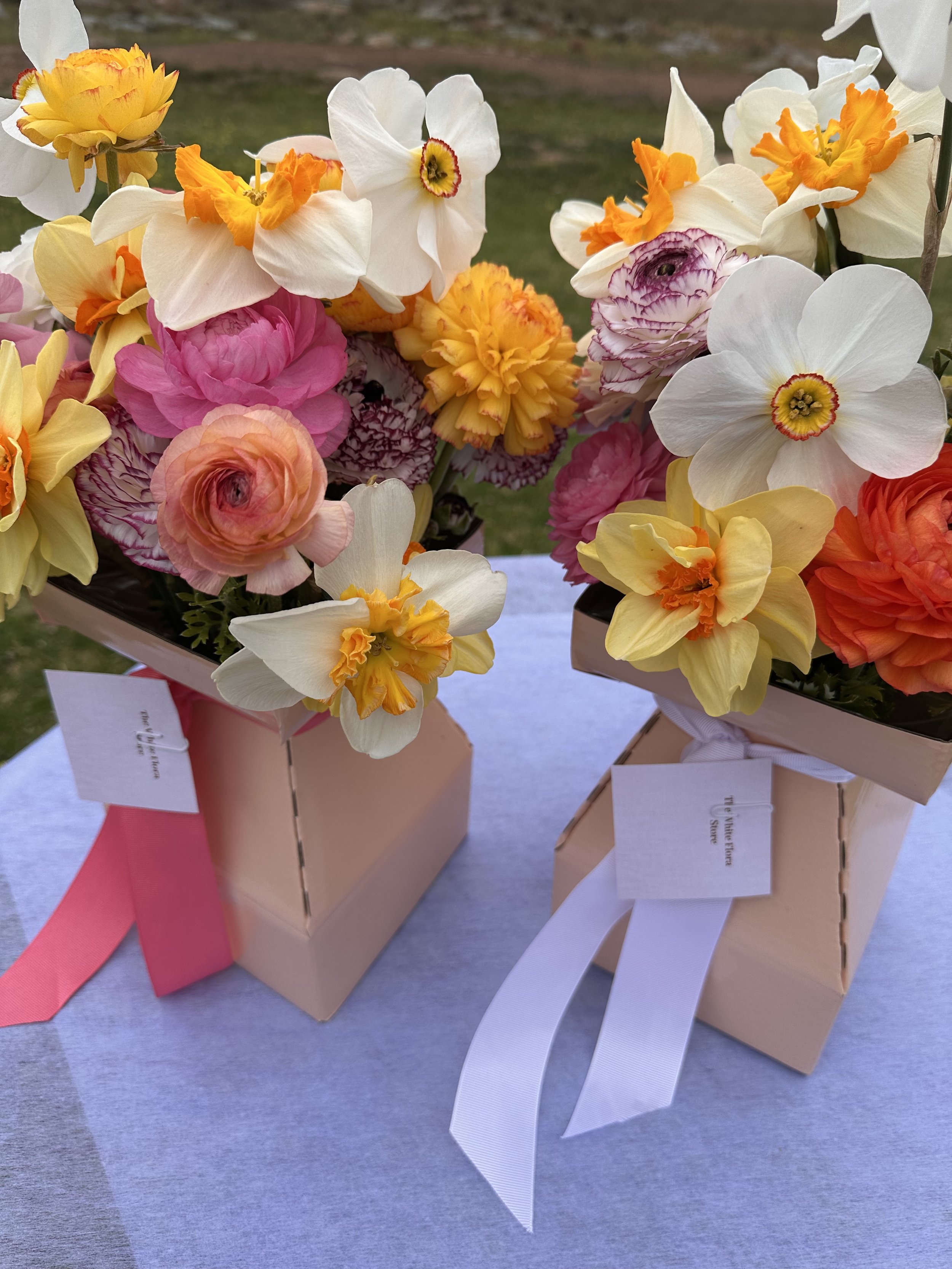 Two pink flower arrangements in pale pink boxes with white and pink ribbons, placed on a surface outdoors.
