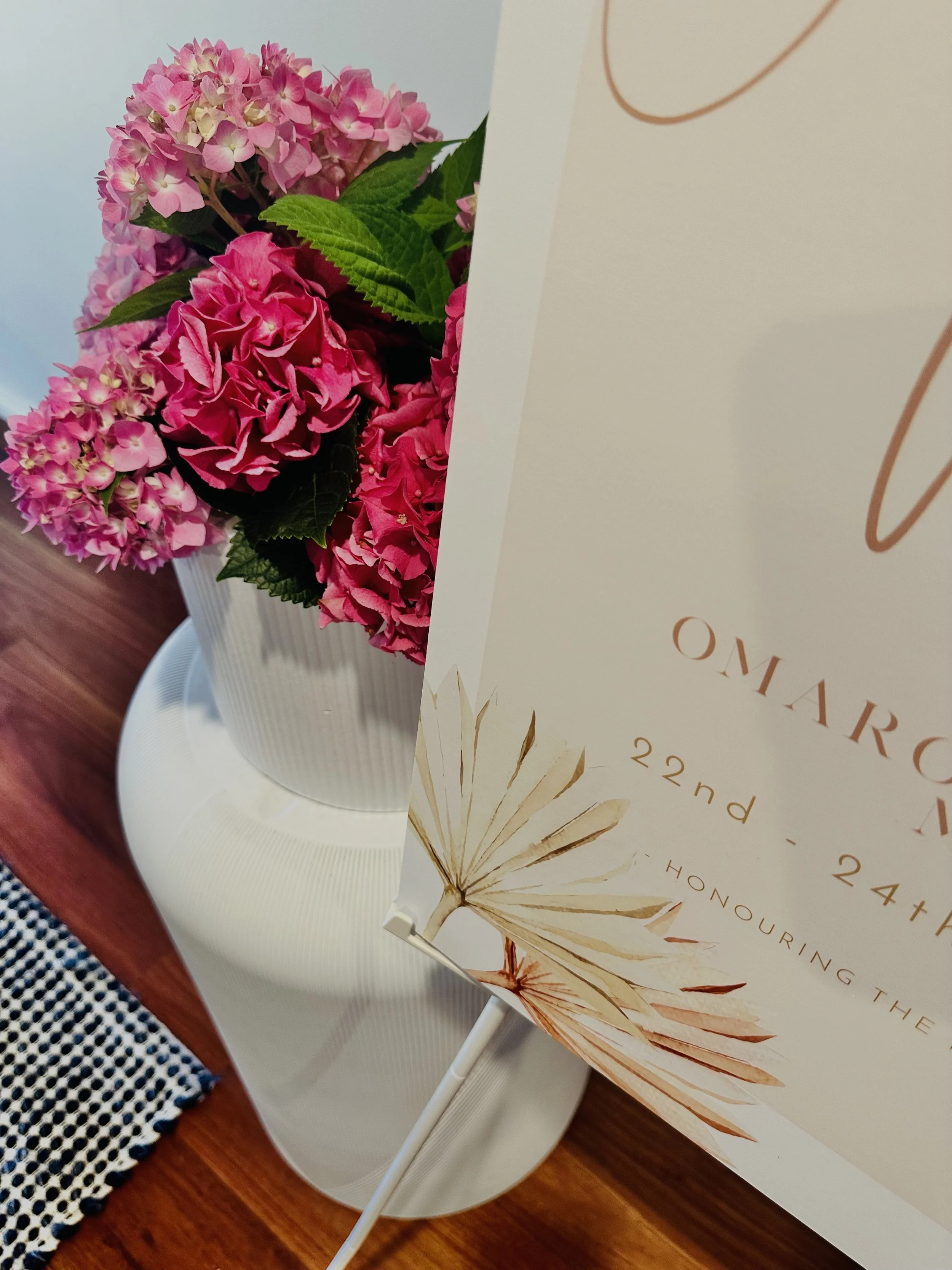 Close-up of pink and purple hydrangea flowers in a striped white vase on a round white table, with a event flyer featuring a beige dried leaf design and partially visible text overlaying the image.