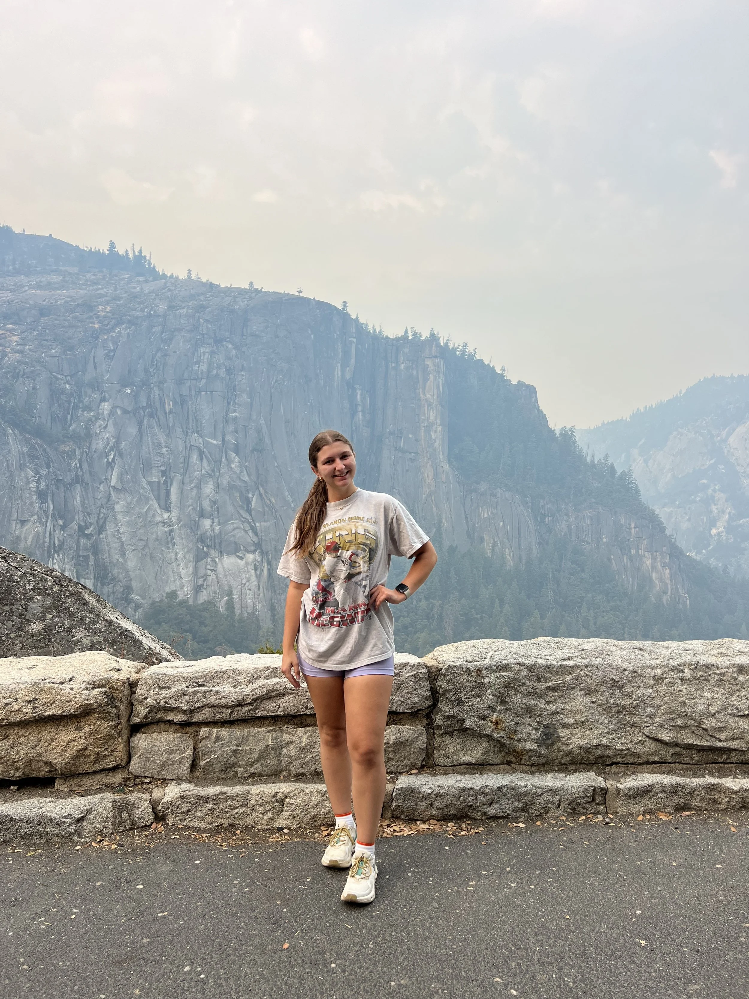 A young woman smiling and posing in front of a mountain landscape with rocky cliffs and pine trees, wearing a t-shirt, shorts, sneakers, and a watch.