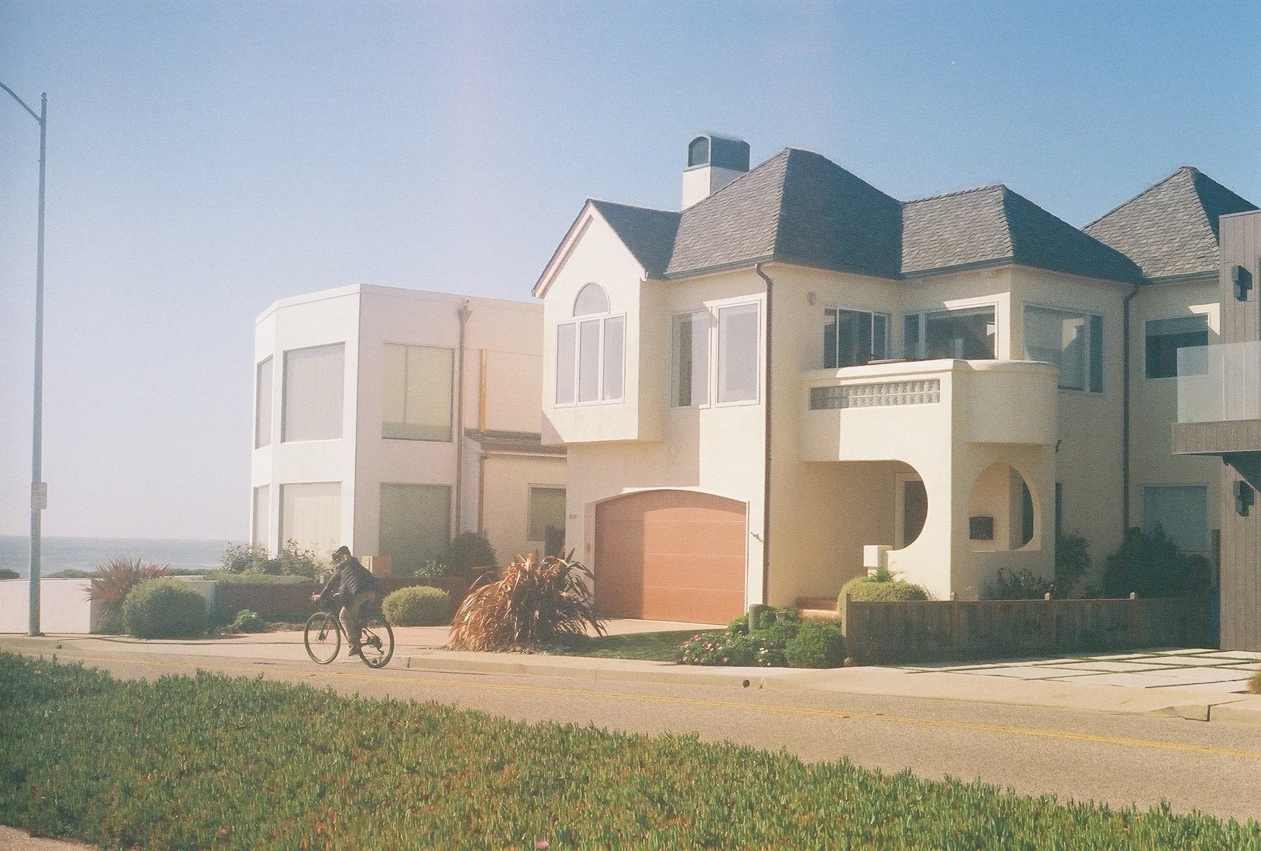 A coastal residential neighborhood with modern houses, a cyclist on the sidewalk, and greenery in the foreground. The sky is clear and blue.