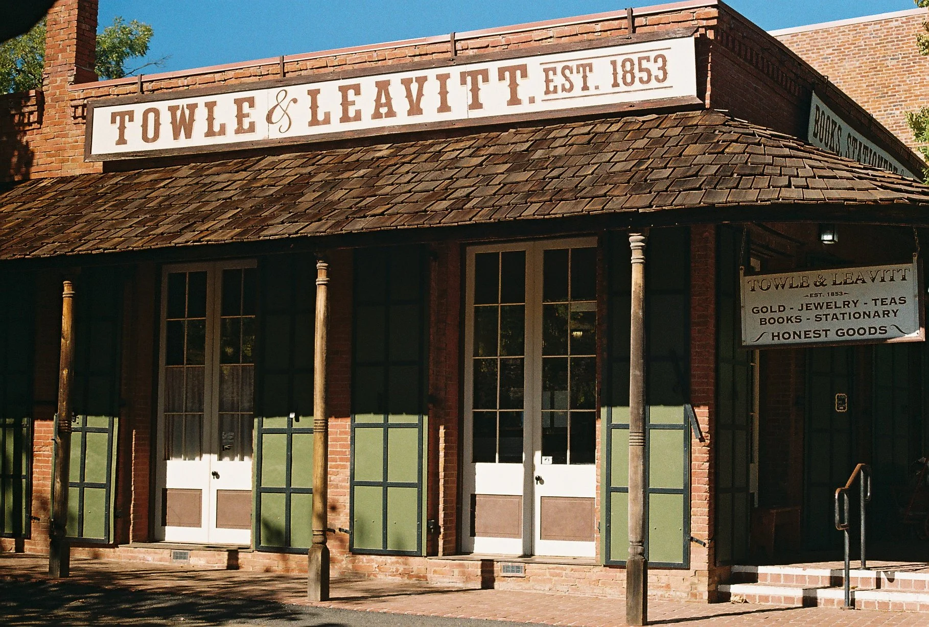 Exterior of a historic brick building with a sign reading "TOWLE & LEAVITT EST. 1853" and a smaller sign listing items sold: gold, jewelry, teas, books, stationary, honest goods.