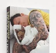 A man with tattoos lying in bed, hugging a dog with a white patch on its neck.