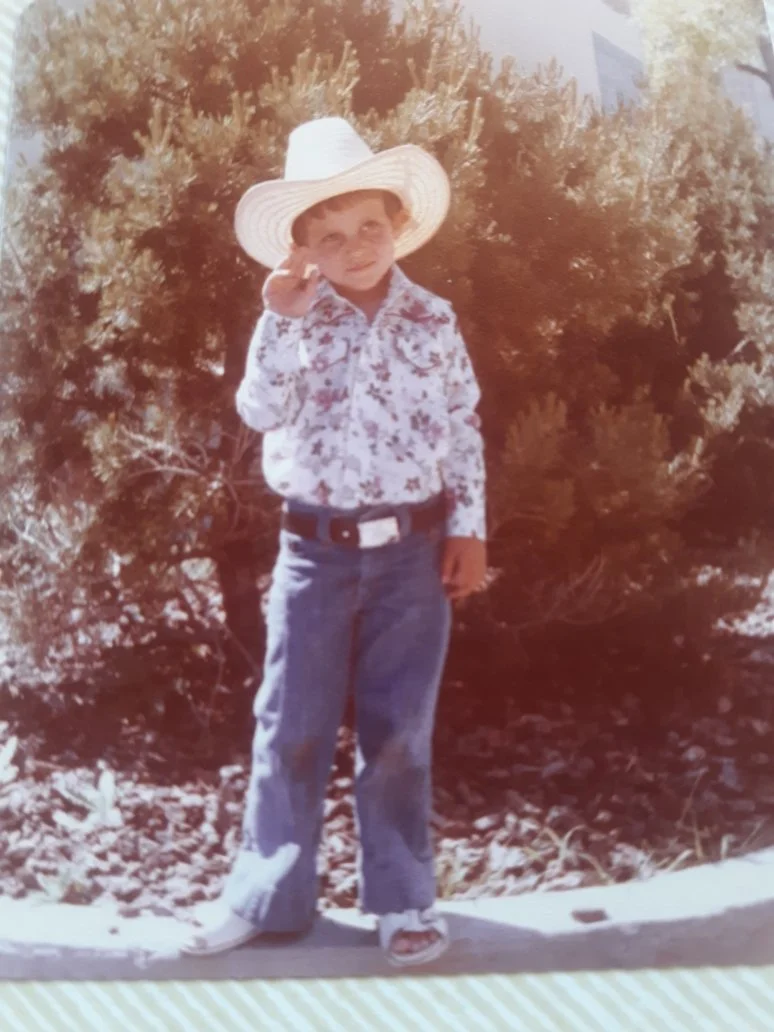 A young boy wearing a cowboy hat, floral shirt, jeans, and sneakers standing outdoors in front of bushes, saluting or shielding his eyes from the sun.