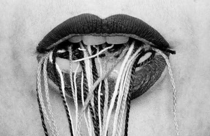 Close-up of a person's mouth with black lipstick, tangled yarn and thread inside the mouth, resembling a messy, yarn-filled tongue.