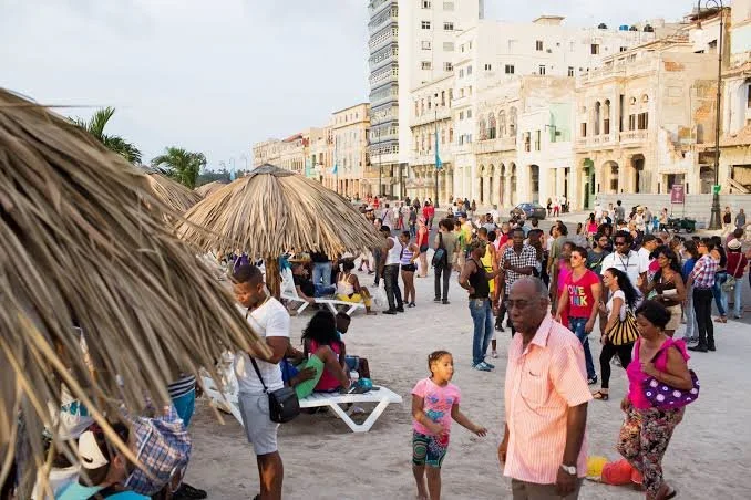 People walking and socializing on a busy beach promenade with old and modern buildings, some under straw thatched umbrellas.