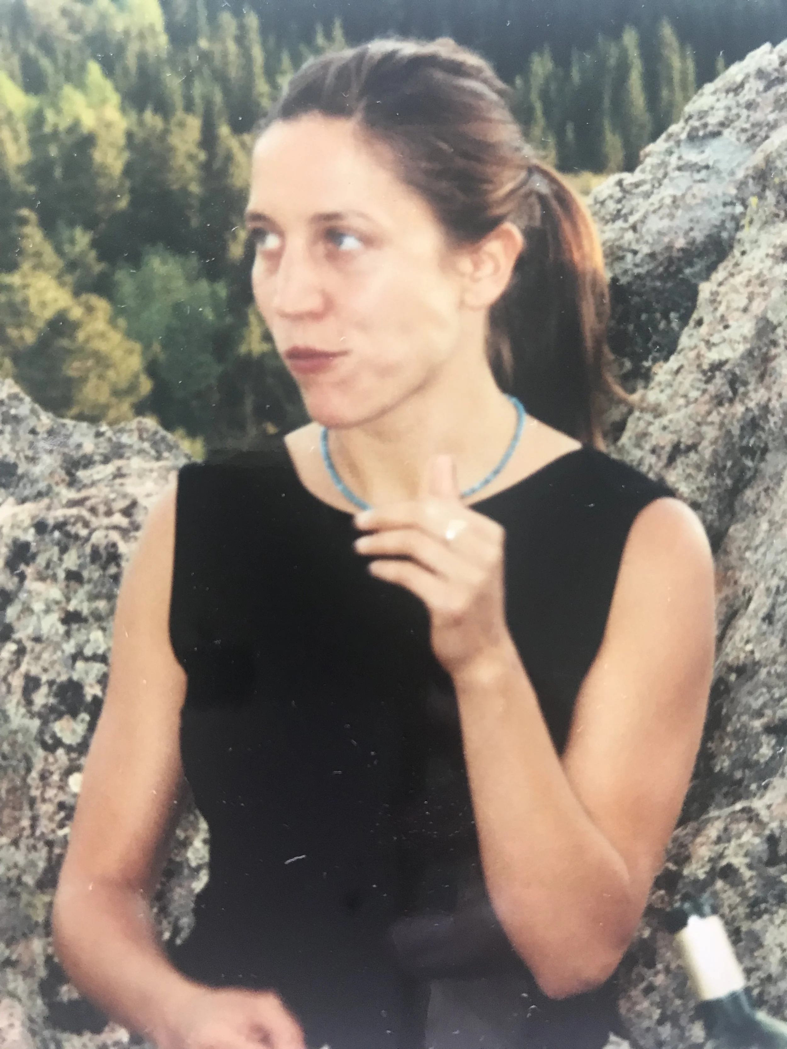 A woman with brown hair tied back, wearing a black sleeveless top, sitting among rocks outdoors with a forested background, holding a glass of wine.