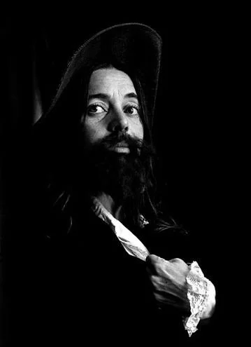 A man dressed as a historical figure, wearing a wide-brimmed hat, with a long beard, holding a feather quill pen.
