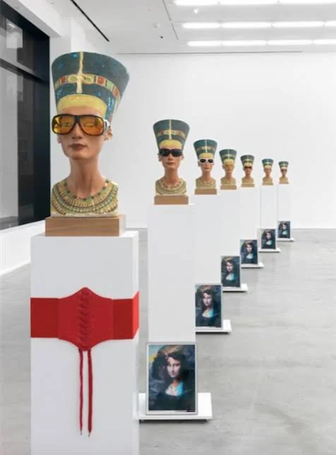 Art exhibition featuring busts of a woman wearing ancient Egyptian-style headdresses and modern sunglasses, displayed on white pedestals with photos of a woman in front of each, arranged in a line.