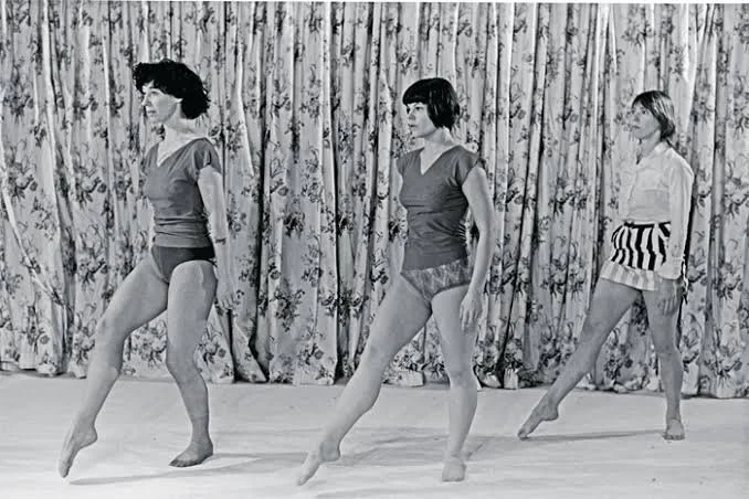 Three women in dance costumes practicing ballet on a wooden floor in front of a floral curtain.