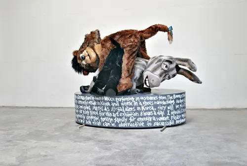 Two large plush toys, a lion and an elephant, are stacked on top of each other on a circular platform with writing on its sides, in a minimalistic gray room.