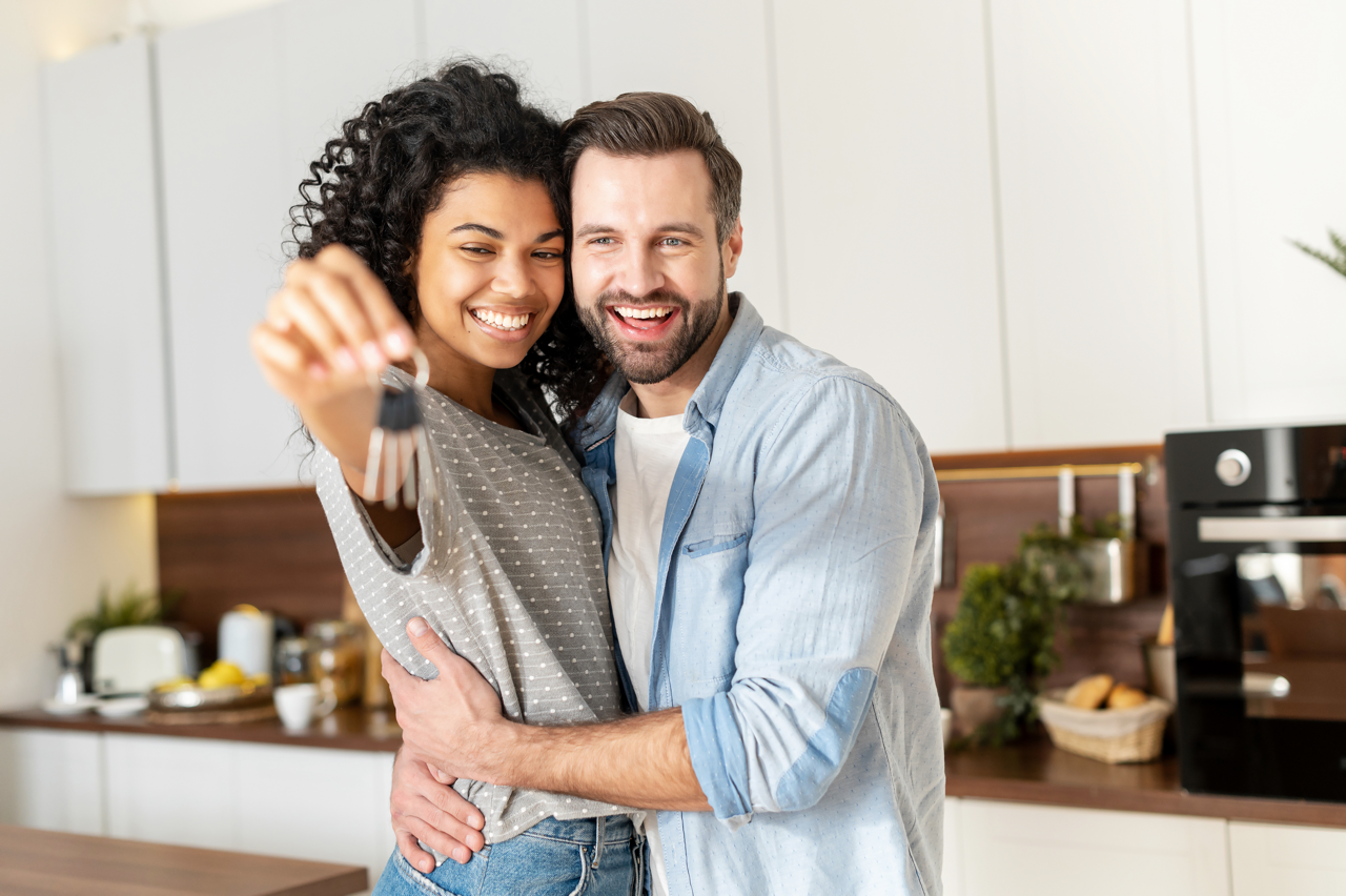 A happy couple smiling in a kitchen, holding a set of keys towards the camera.