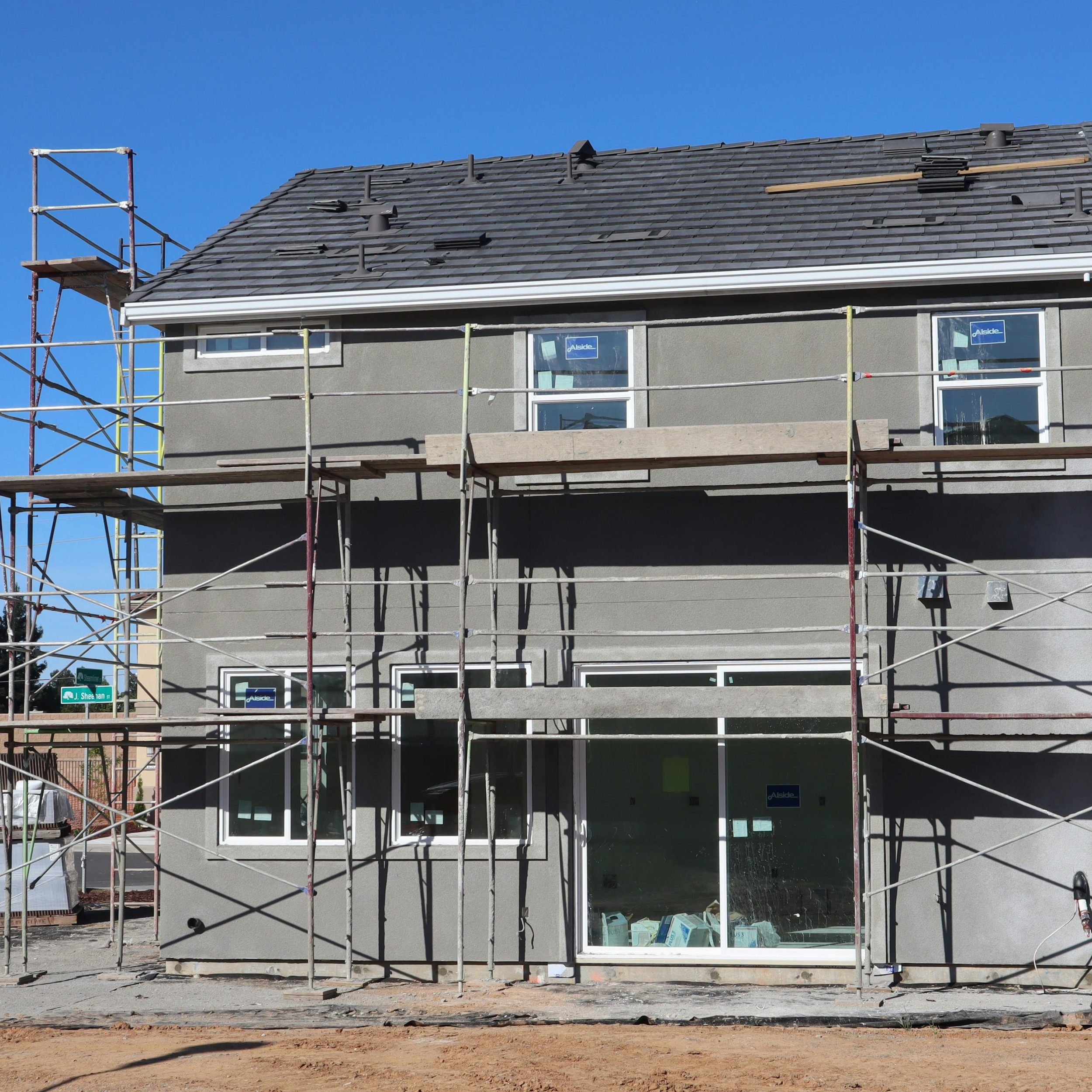 Front view of a house under construction with scaffolding around it, showing three windows and a glass door, and a partly tiled roof against a clear blue sky.